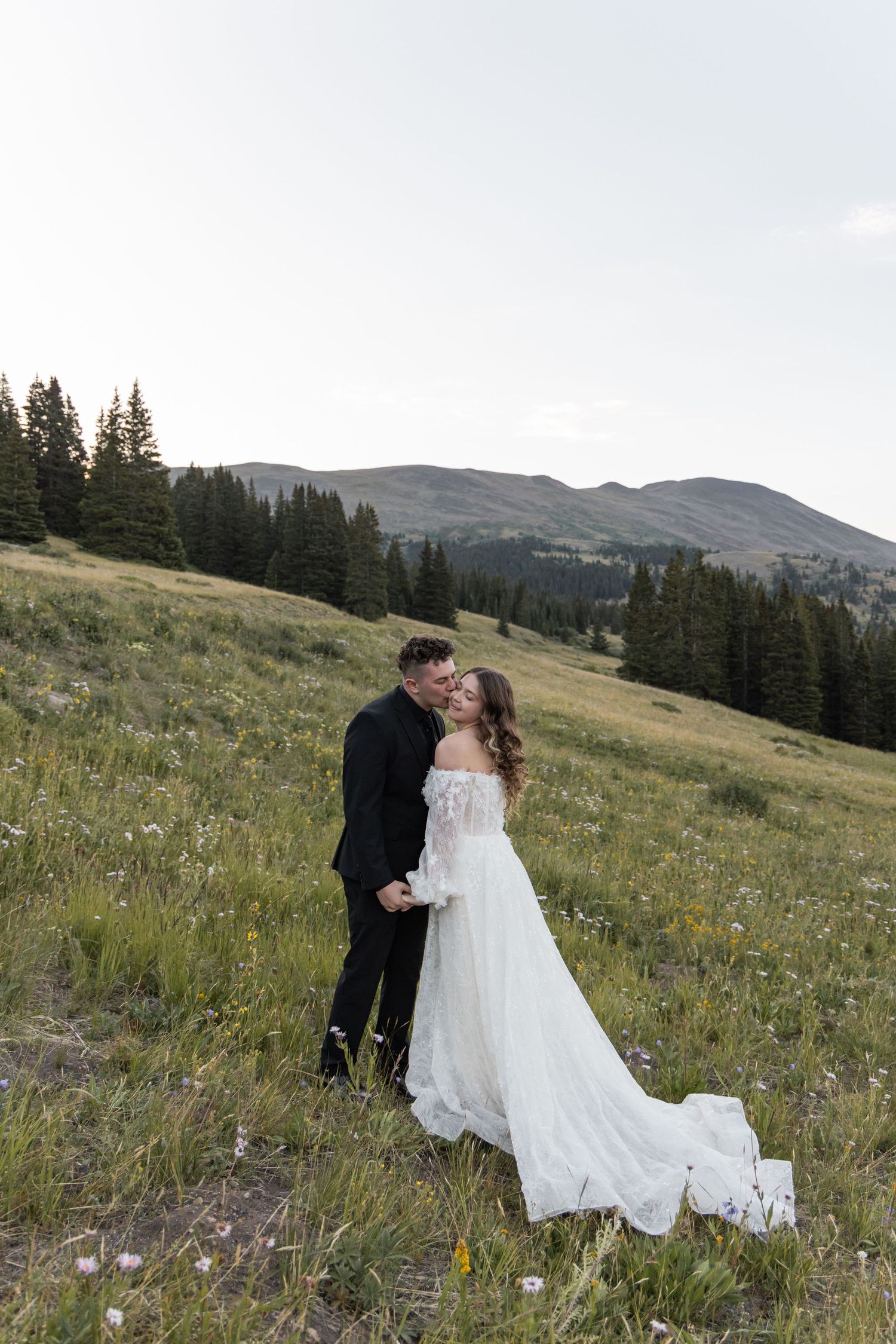 A bride and groom share a romantic kiss in a wildflower meadow with pine trees and mountains in the background.