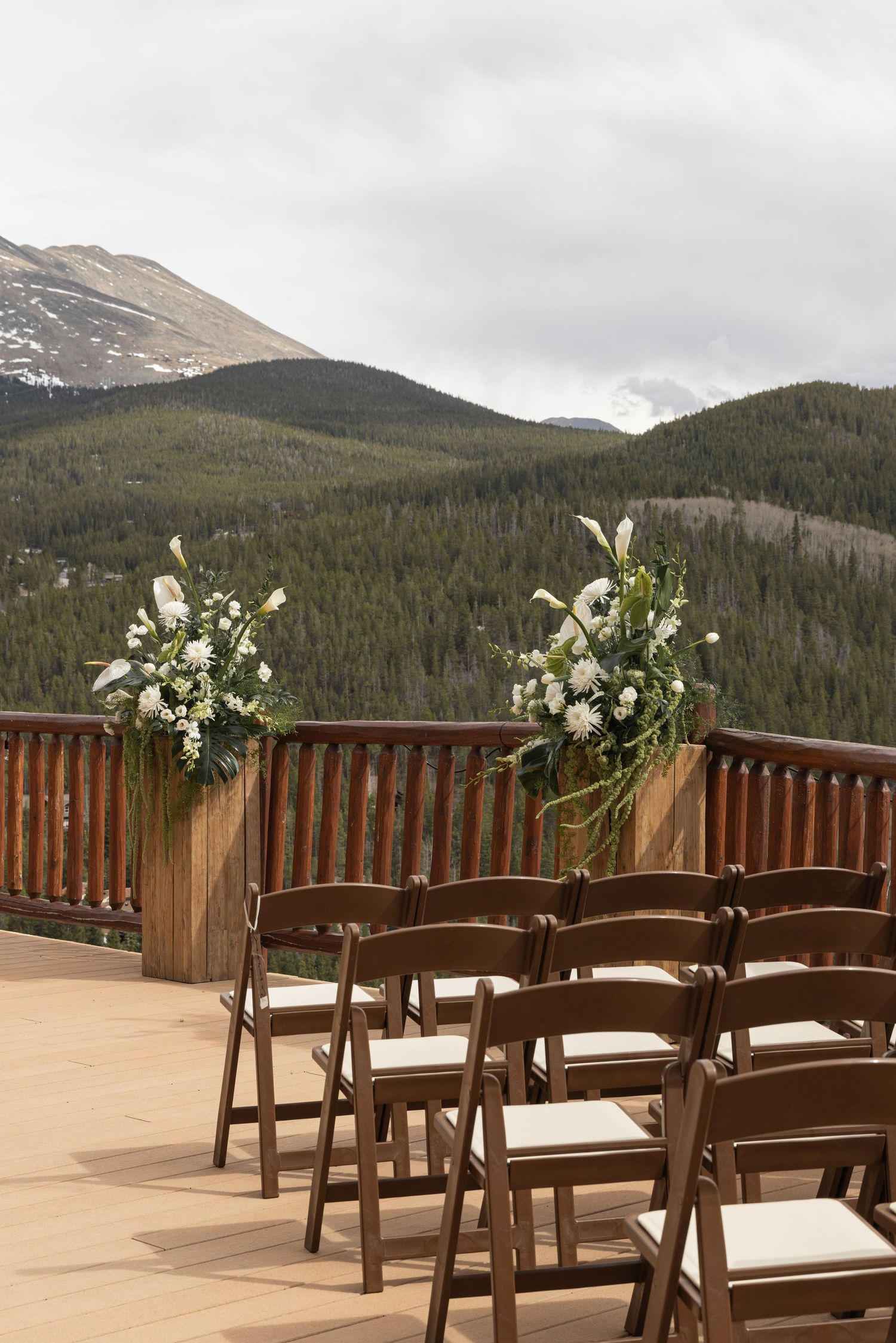 Outdoor mountain wedding ceremony setup with brown folding chairs and white floral arrangements on a wooden deck.