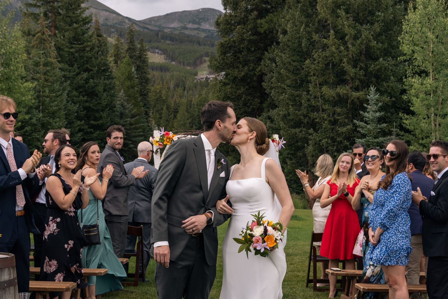 Wedding guests raise champagne glasses in an outdoor mountain celebration with evergreen trees in the background.