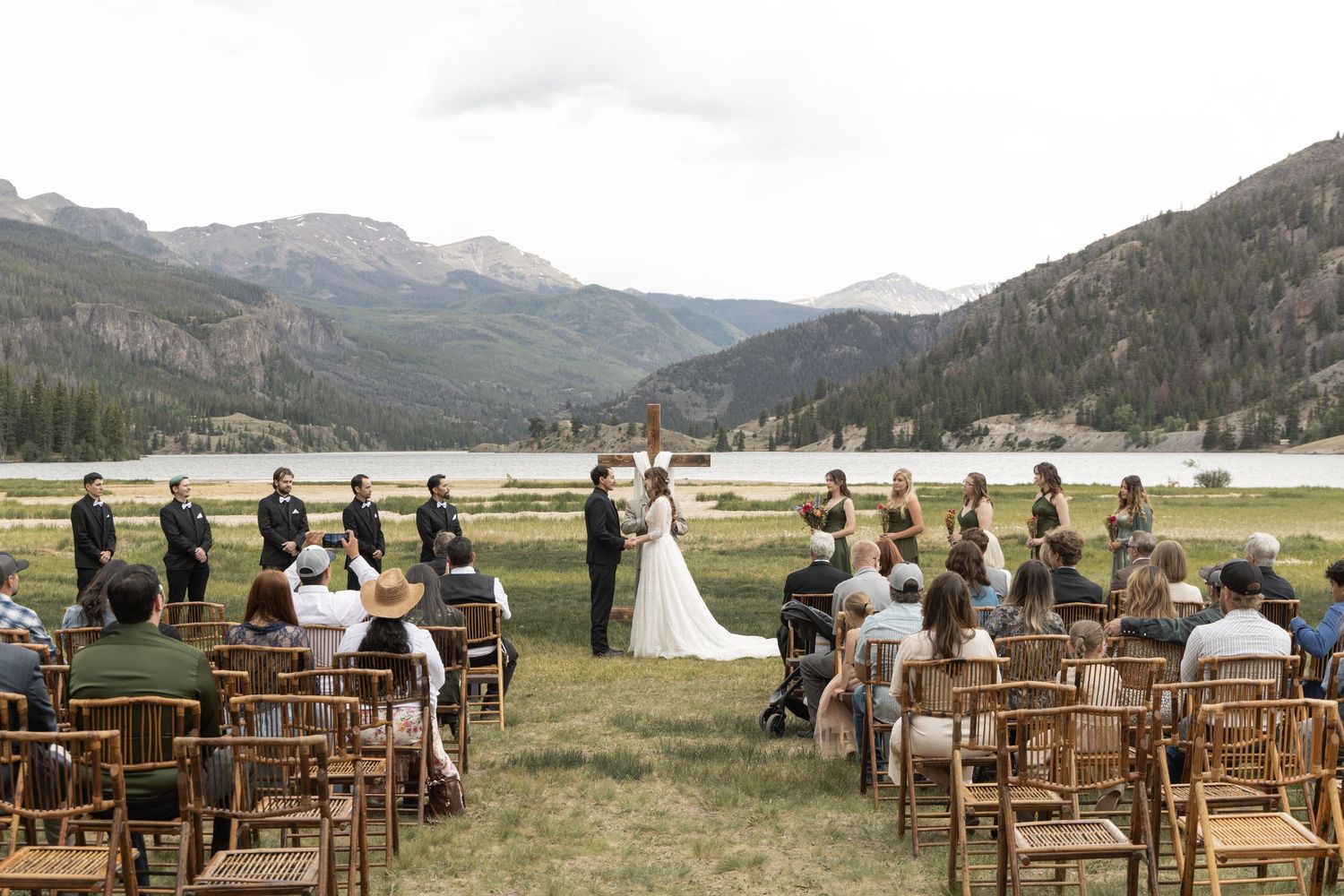 Outdoor mountain wedding ceremony with guests seated in wooden chairs by a scenic lake and rocky peaks.