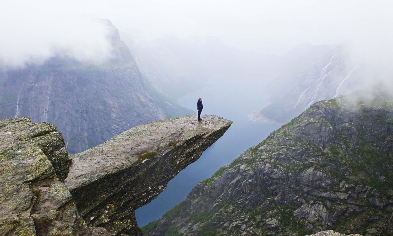 Person standing on Trolltunga rock ledge overlooking misty Norwegian fjord and mountains.