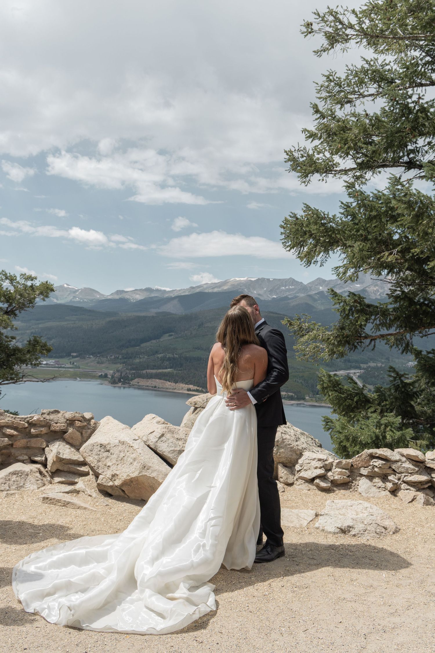 A couple in wedding attire embraces while sitting on a rocky overlook with mountain lake views in the background.