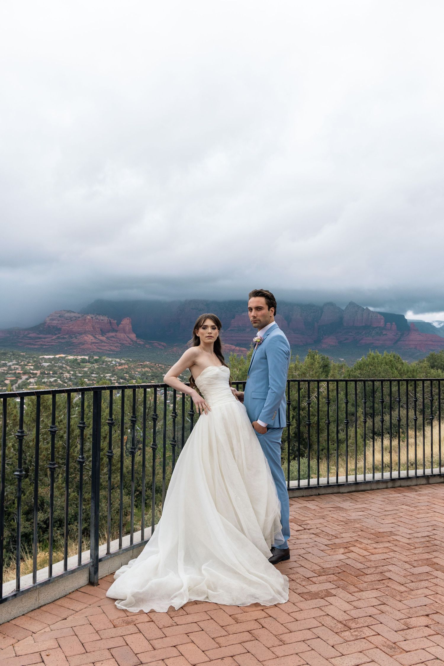 A couple in wedding attire poses on a brick terrace overlooking the scenic red rock mountains of Sedona at sunset.
