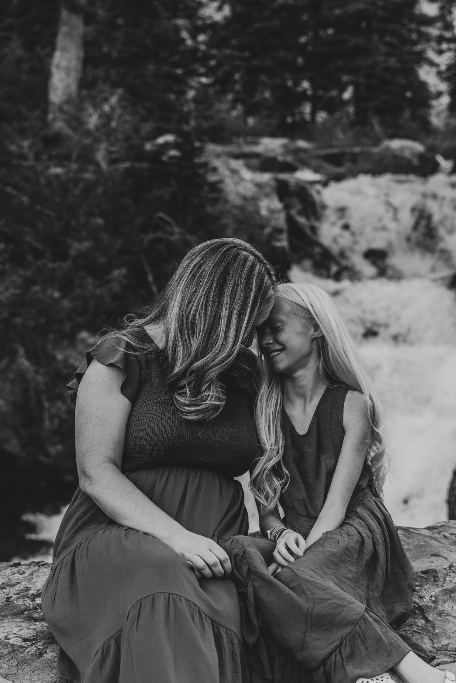 Two people wearing matching dark dresses sit close together on rocks near a forest waterfall in black and white.