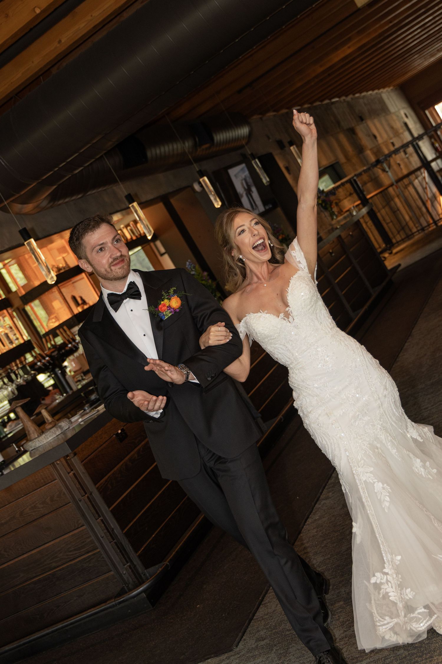 Joyful bride in white lace gown raises arm celebrating with groom in black tuxedo at elegant wedding venue bar.