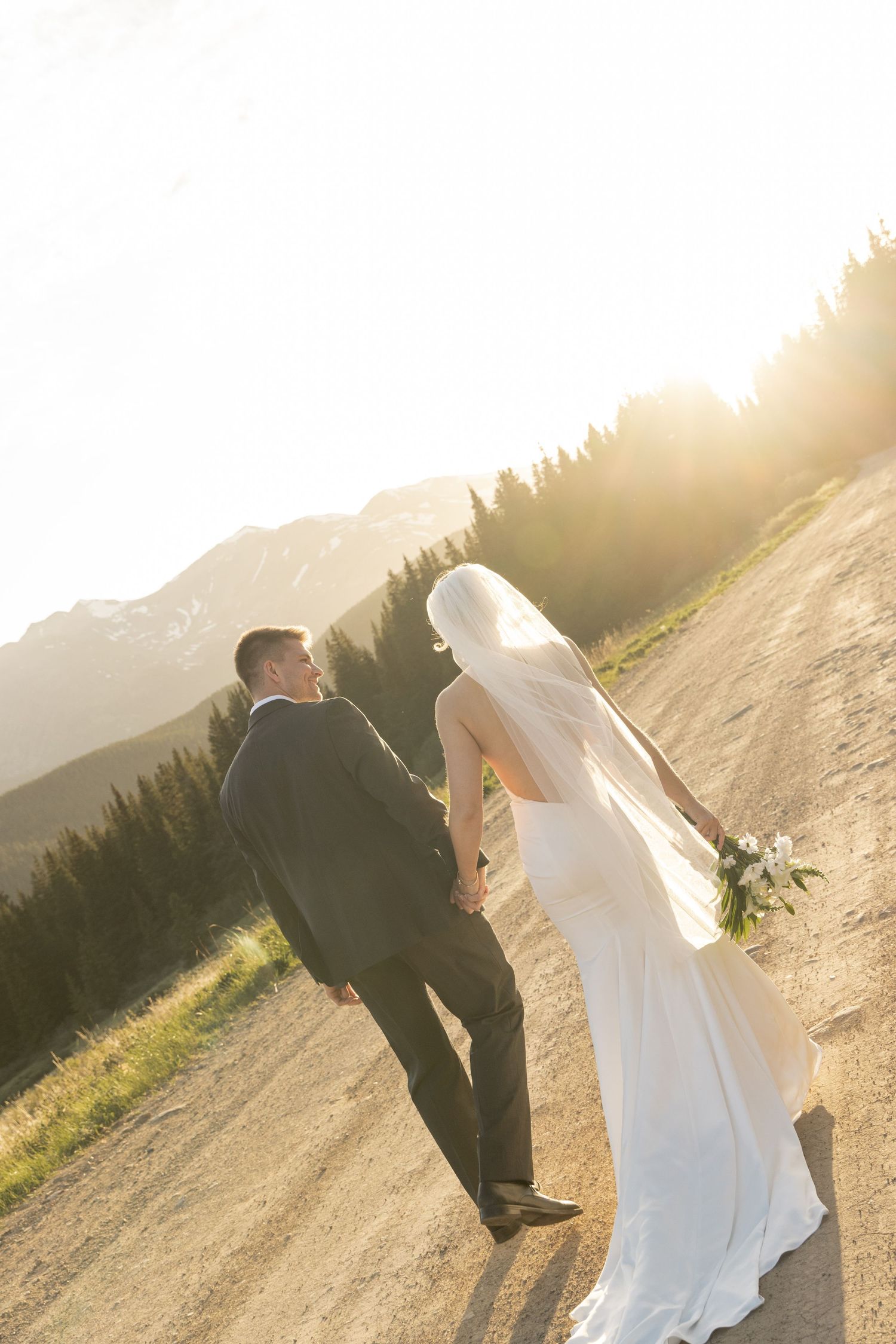 Bride and groom walking hand in hand on a mountain dirt path at golden sunset, with evergreen trees and peaks behind them.