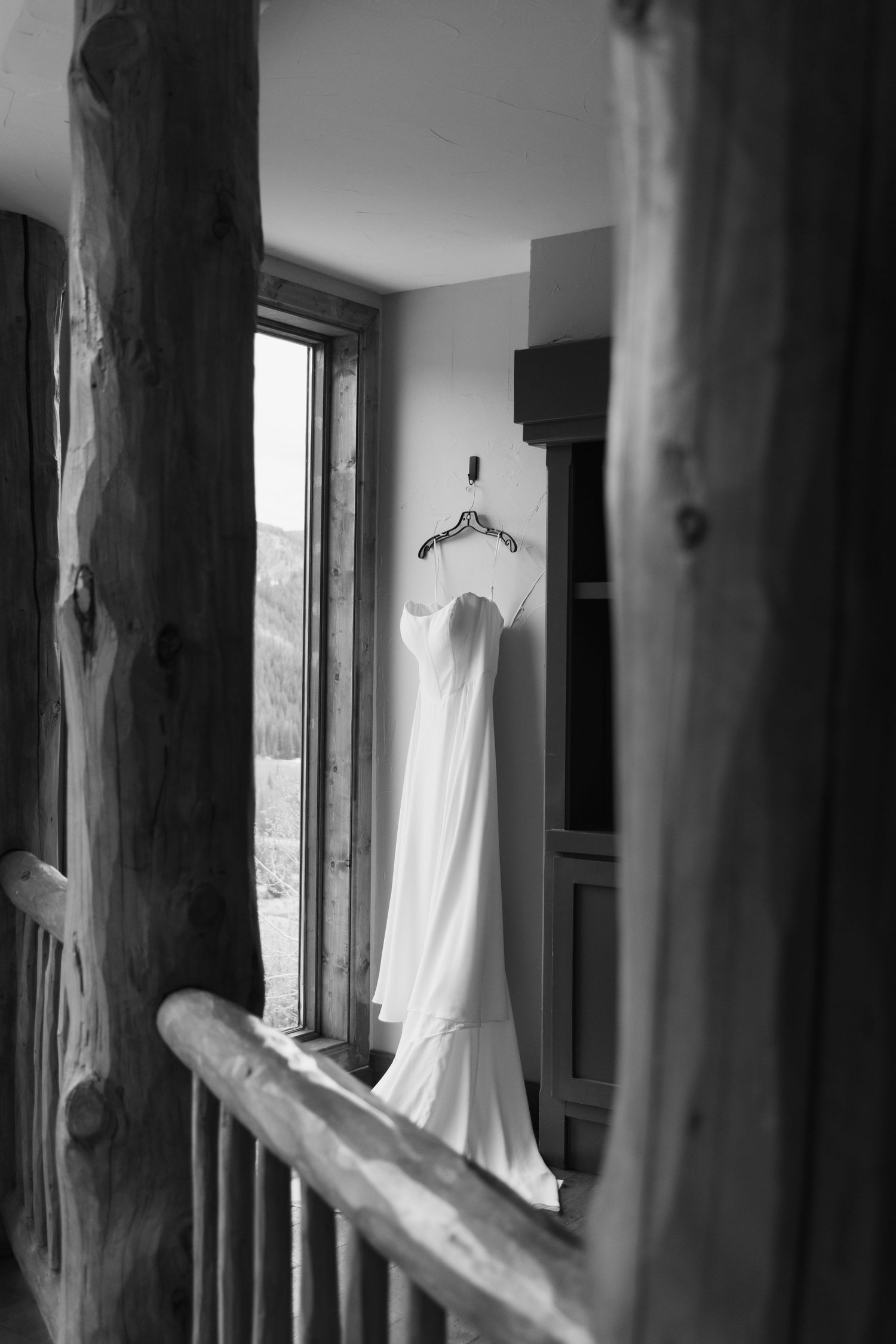Black and white photo of an elegant wedding dress hanging on a hanger near a window in a rustic log cabin.