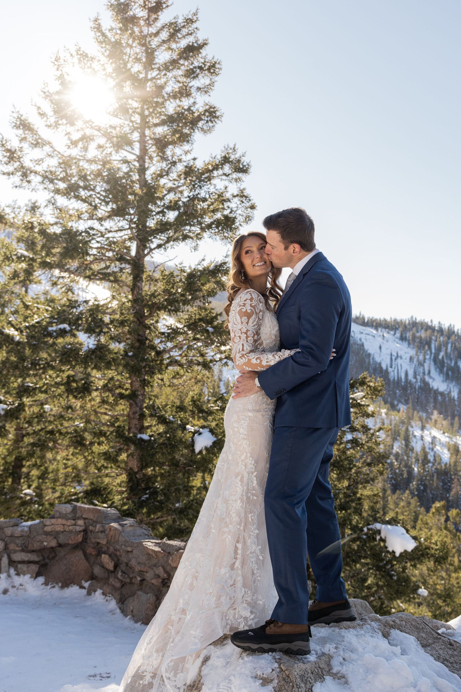 A couple shares an intimate winter wedding moment on a snowy mountain overlook with evergreen trees in the background.