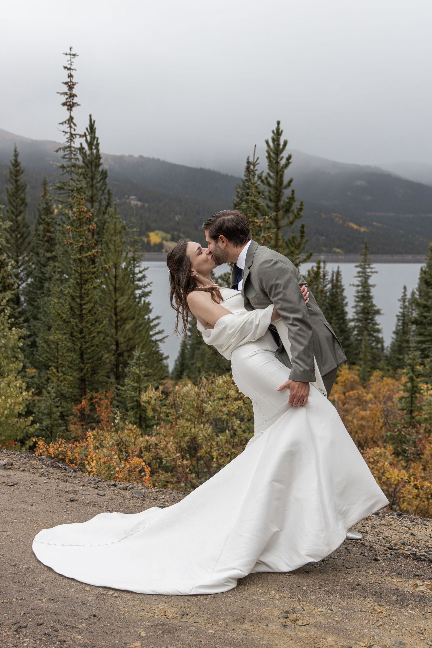 Bride and groom share a romantic dip kiss surrounded by pine trees and a mountain lake on their wedding day.