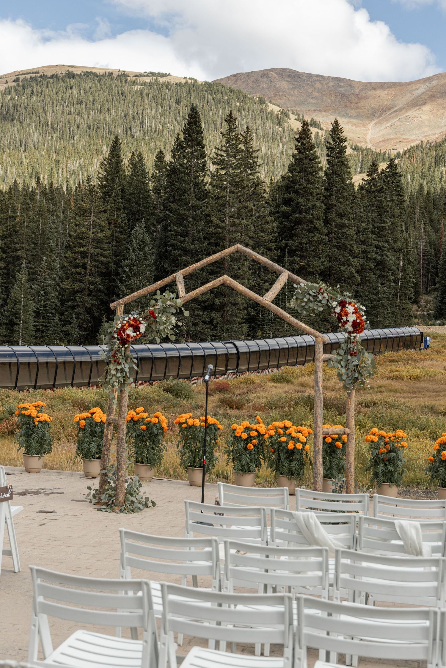 White chairs arranged for an outdoor mountain wedding ceremony with rustic wooden arch decorated with autumn flowers.