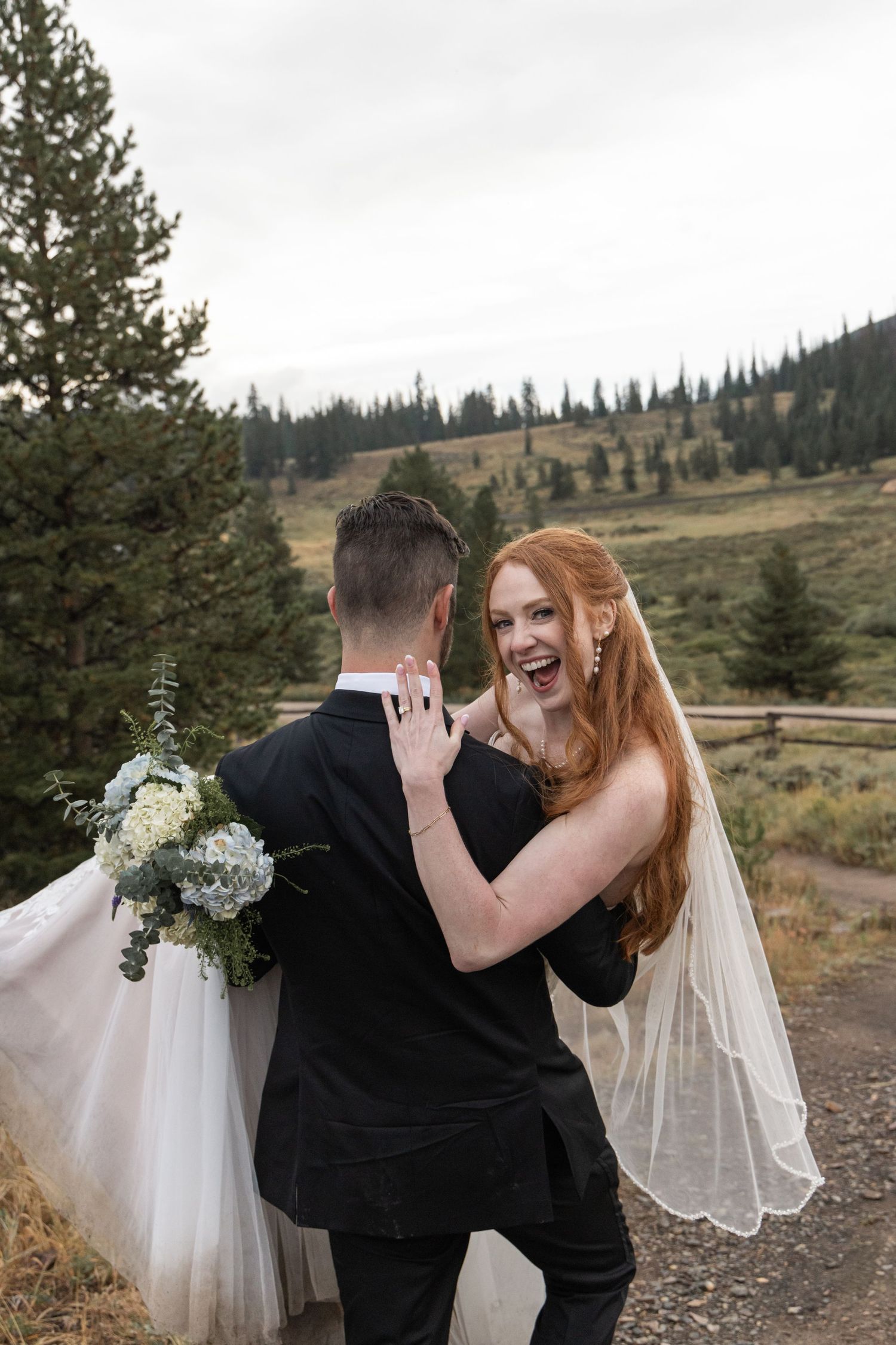 Joyful bride with red hair and veil laughs with groom holding bouquet, outdoor mountain wedding setting.