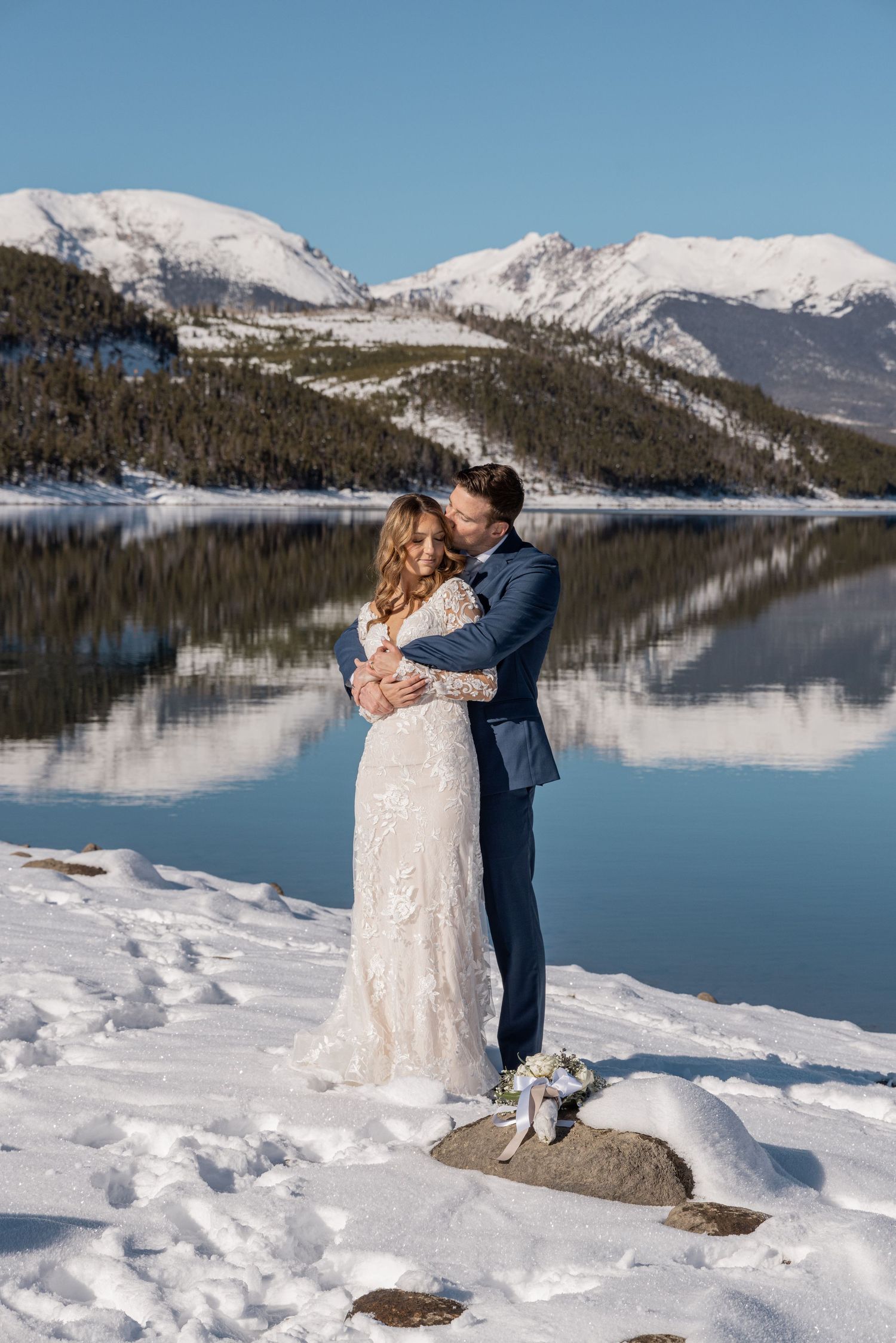 Bride and groom embracing by a snowy mountain lake, wearing wedding attire, with snow-capped peaks reflecting in calm water.