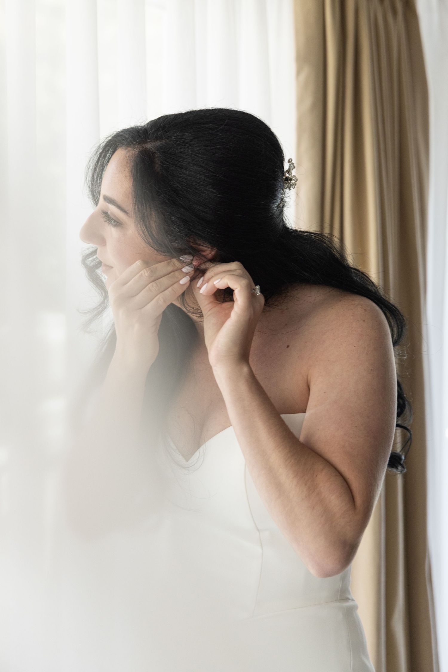 Bride in white dress putting on earrings by a window with sheer curtains on her wedding day.