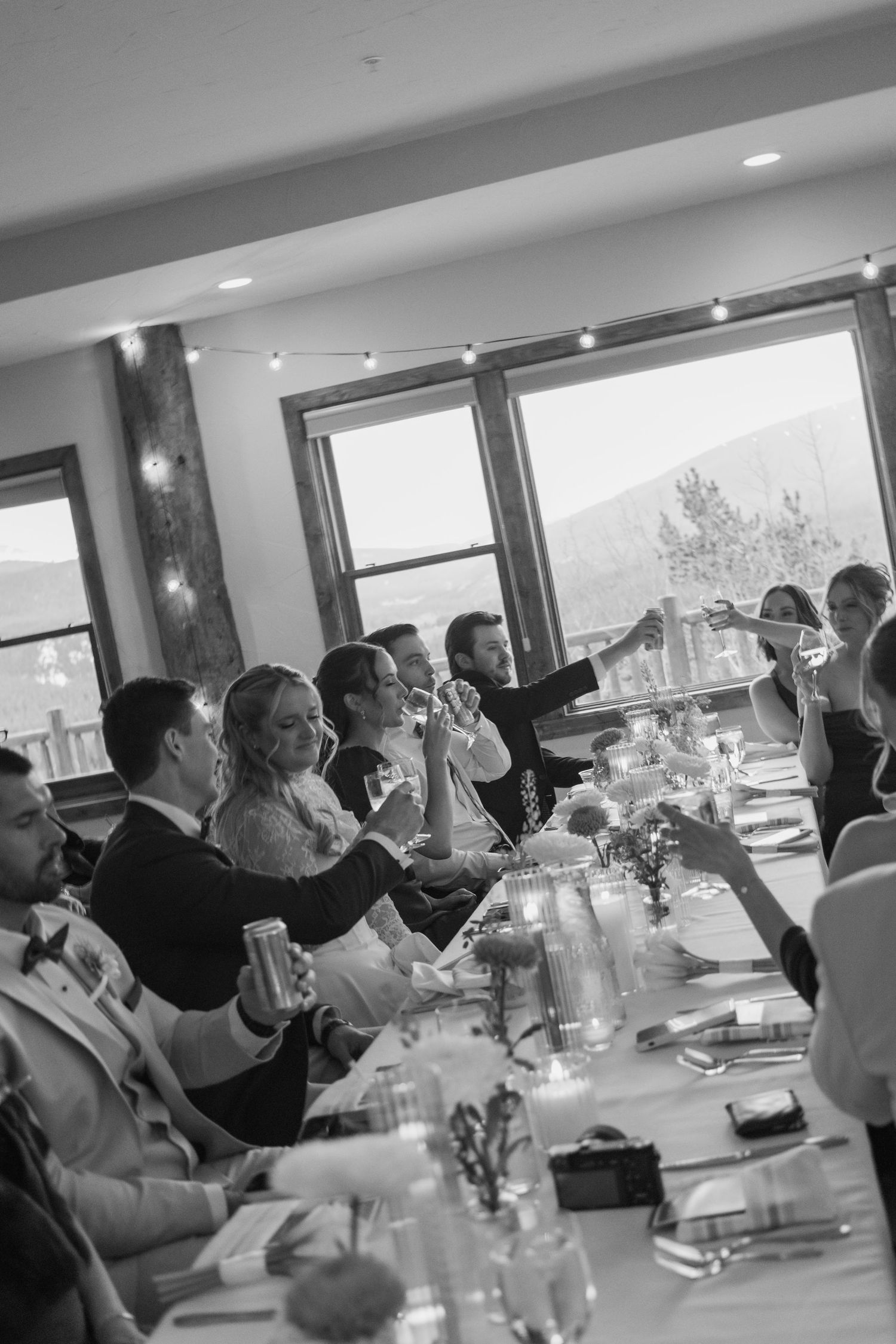 Black and white wedding reception photo of guests raising glasses in a toast at a long decorated table with mountain views.