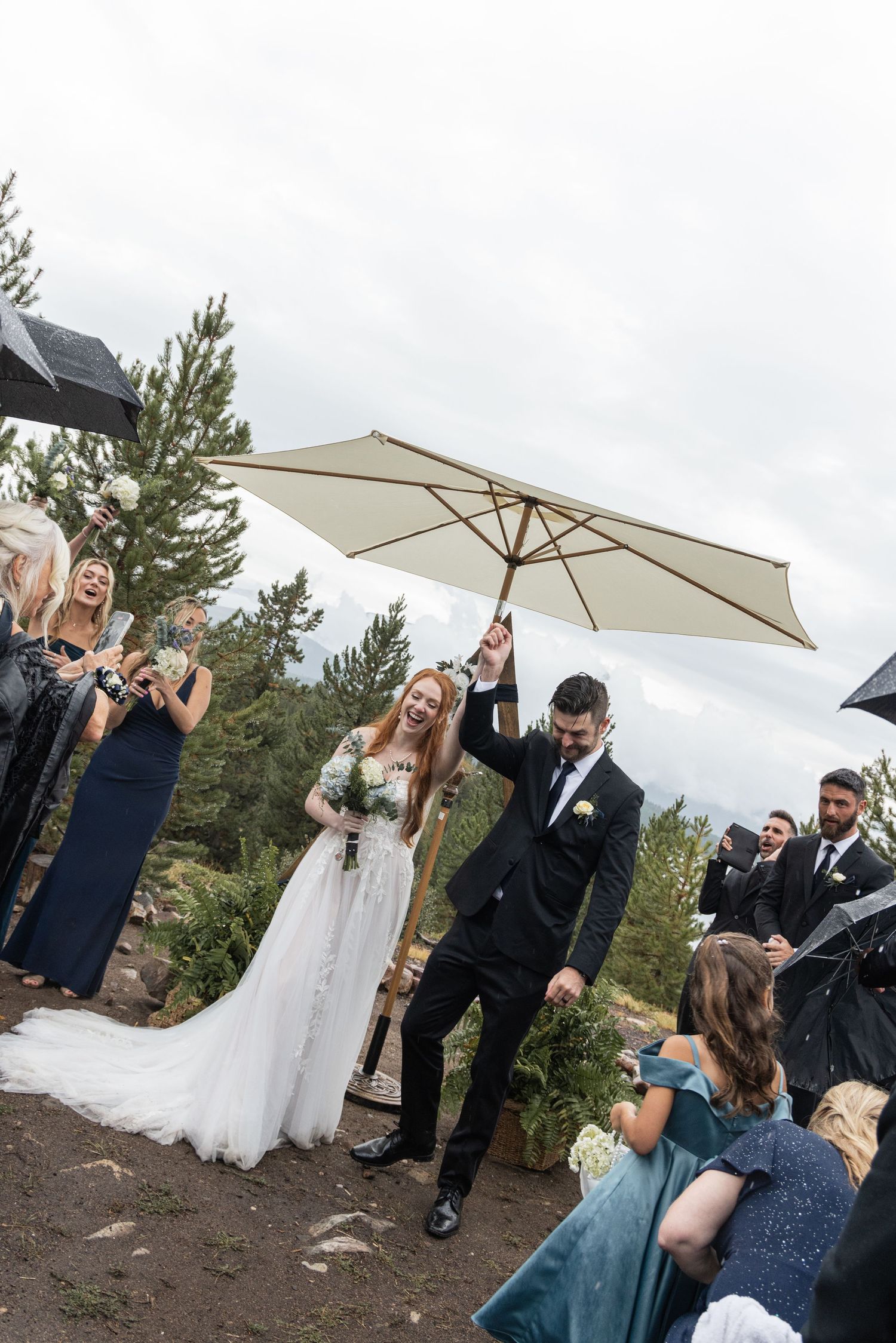 Wedding ceremony outdoors in nature with white umbrella and pine trees in background during cloudy weather.