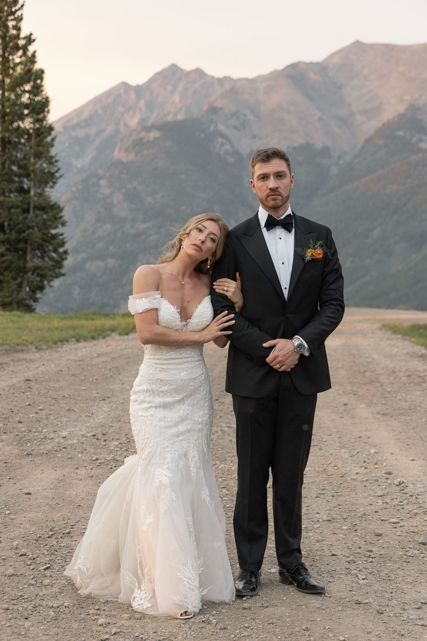 Bride in off-shoulder lace gown and groom in black tuxedo pose on mountain gravel road at sunset.