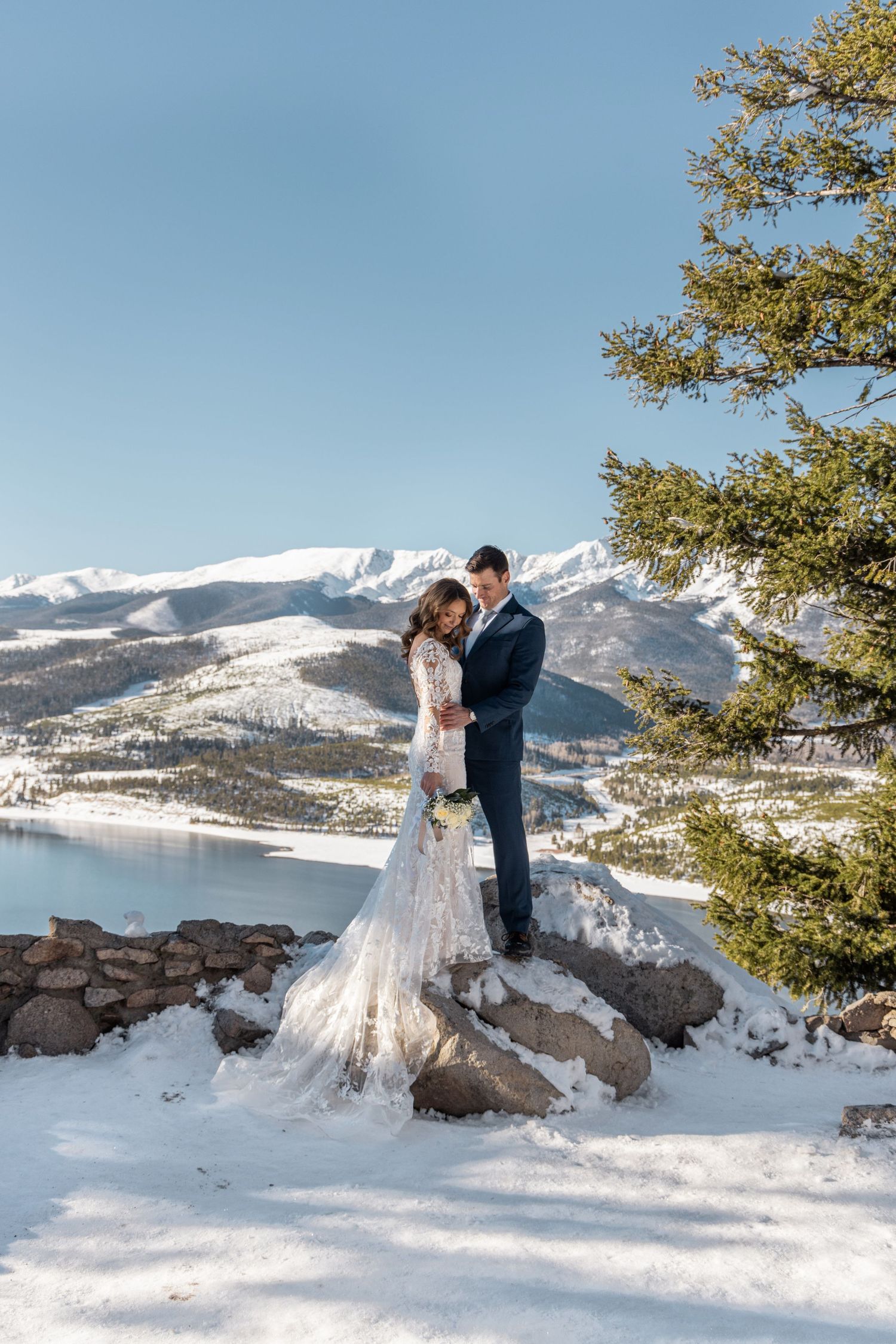 A couple embraces on a snow-covered mountaintop with lake and snowy peaks in the backdrop.