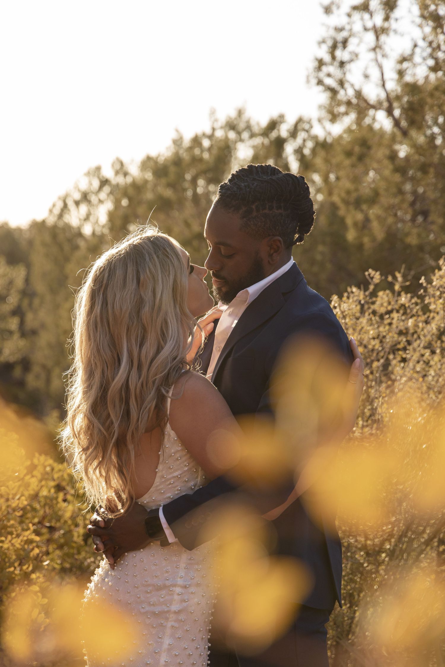 A romantic wedding couple embraces in a golden field at sunset with soft bokeh and wildflowers in the foreground.
