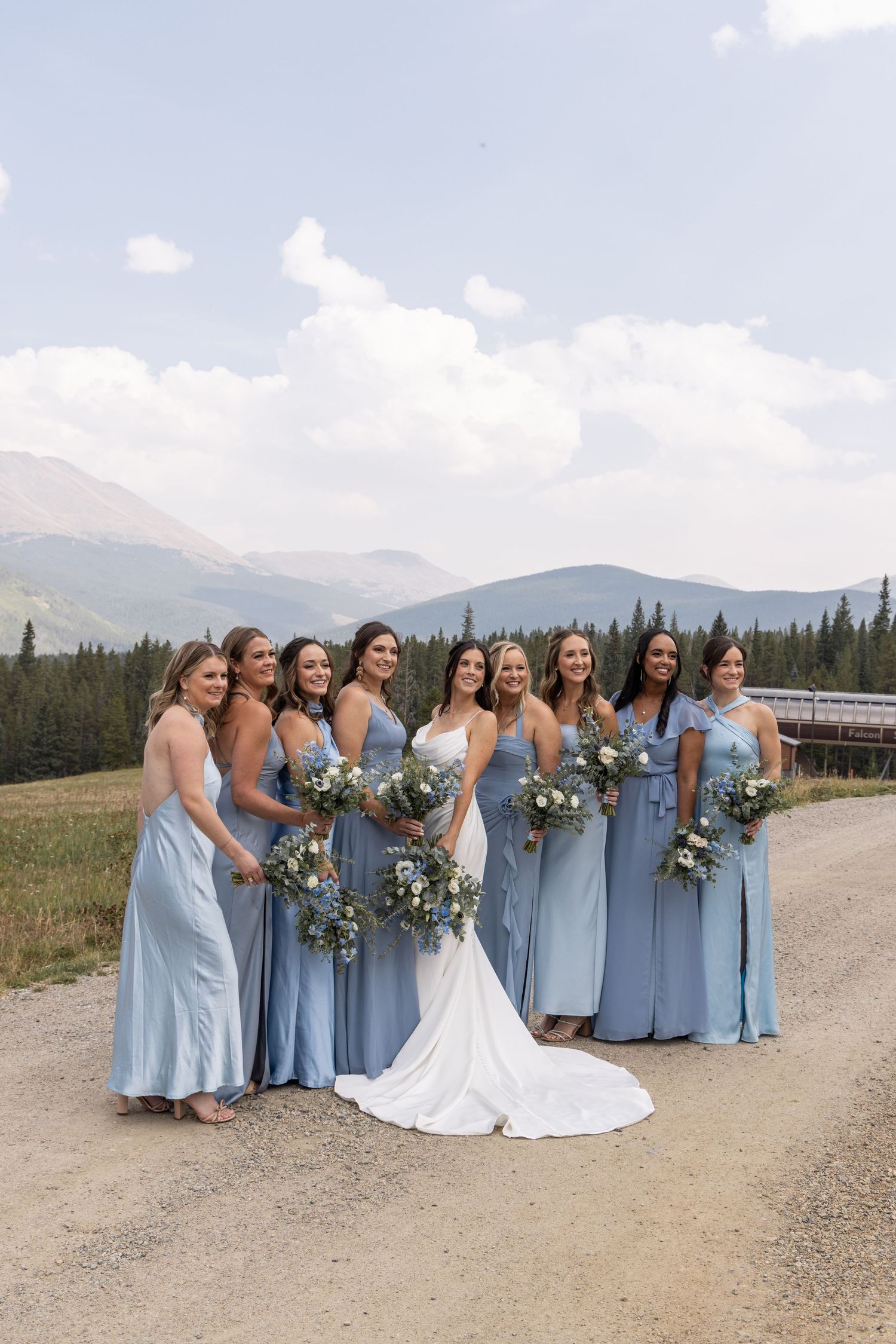 Bride in white gown with bridesmaids in blue dresses holding bouquets, posing outdoors with mountain backdrop.