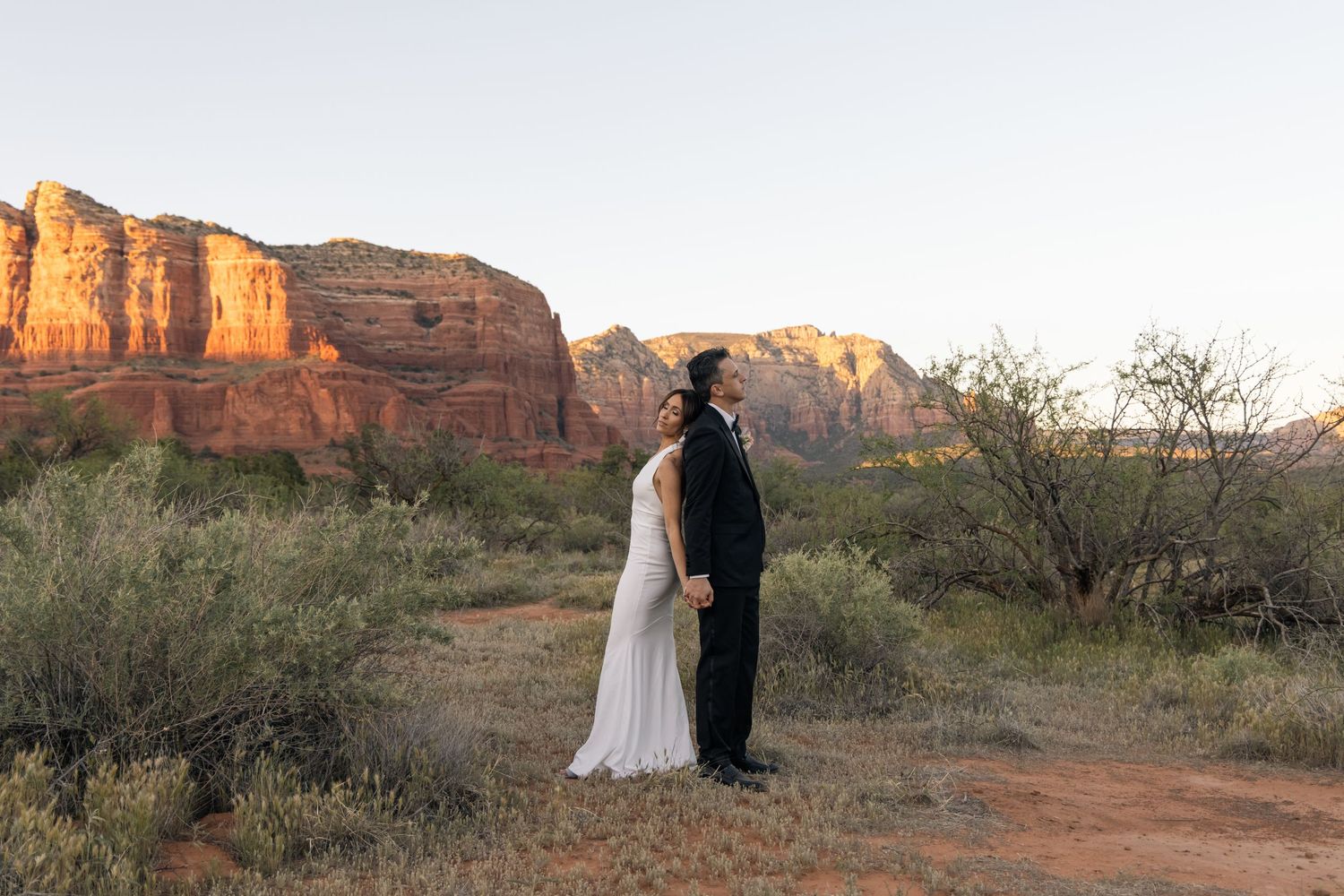Couple in wedding attire standing together in Sedona Arizona desert landscape with red rock formations at sunset.
