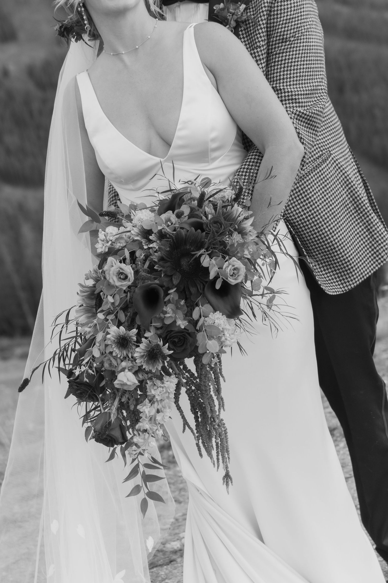 Black and white photo of a bride in a deep V-neck gown holding a cascading floral bouquet with sunflowers and roses.