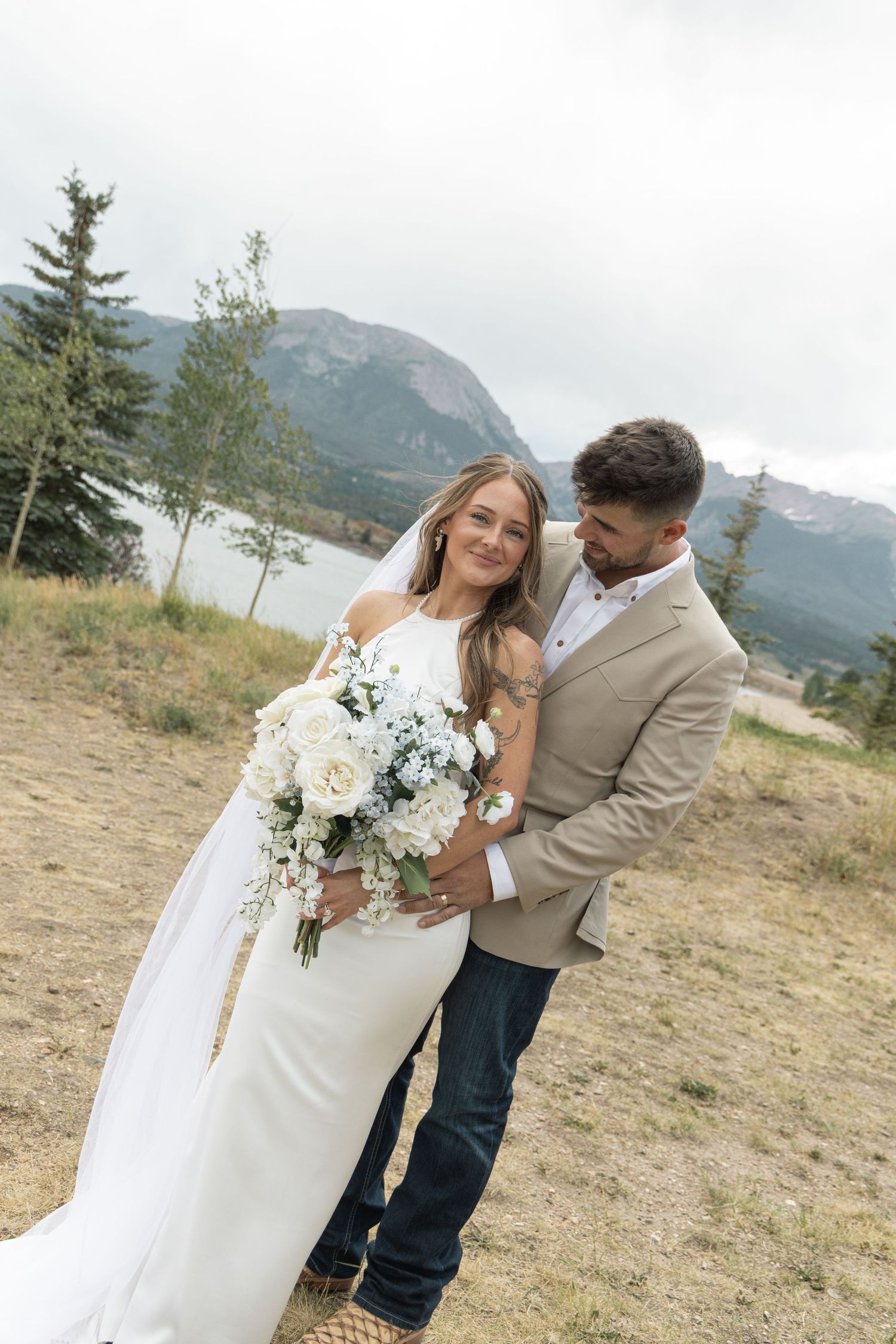 Bride and groom embrace outdoors near a mountain lake, she holds a white bouquet, wearing a white gown and veil.