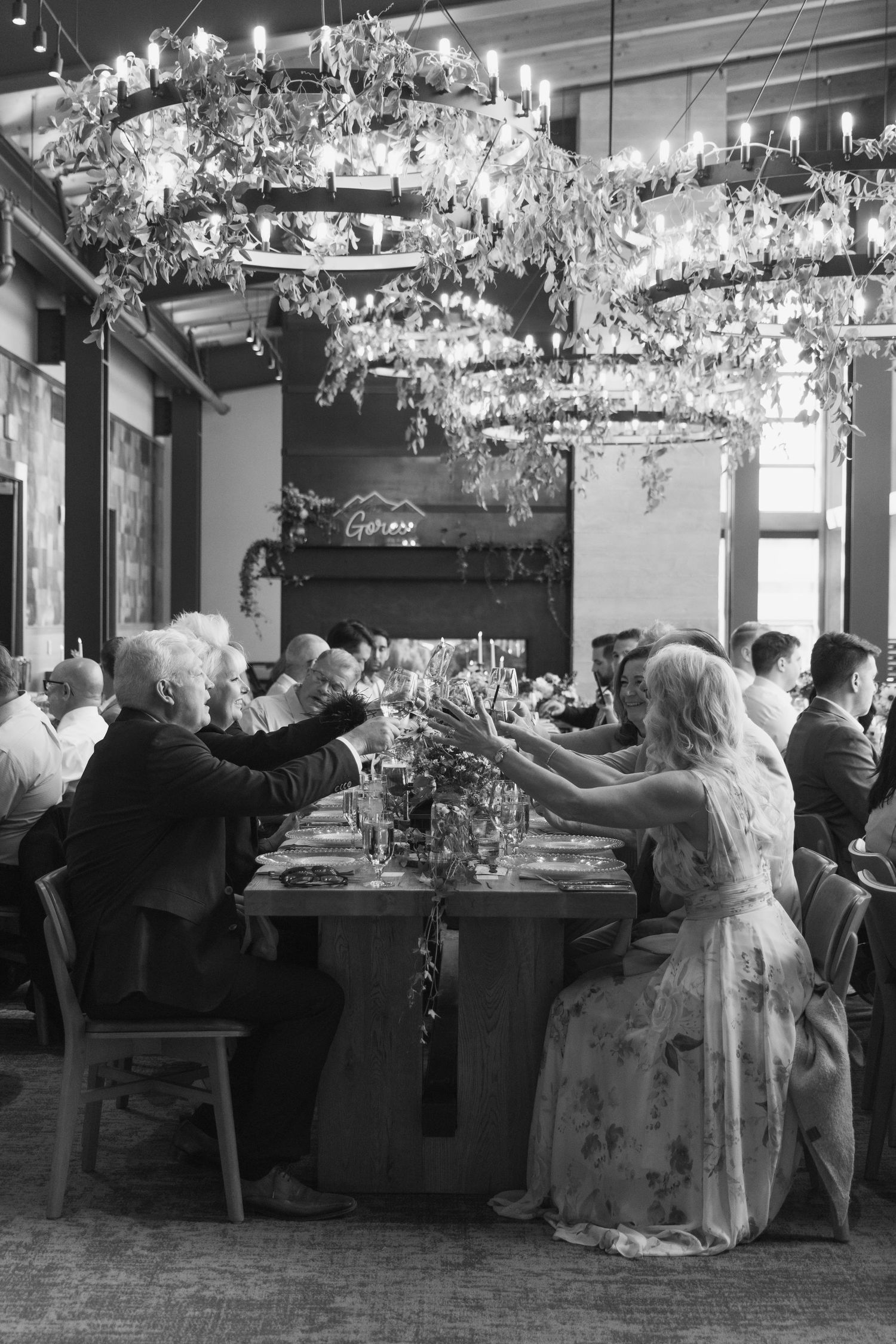 Black and white photo of wedding reception guests toasting at a long decorated table under floral chandeliers.