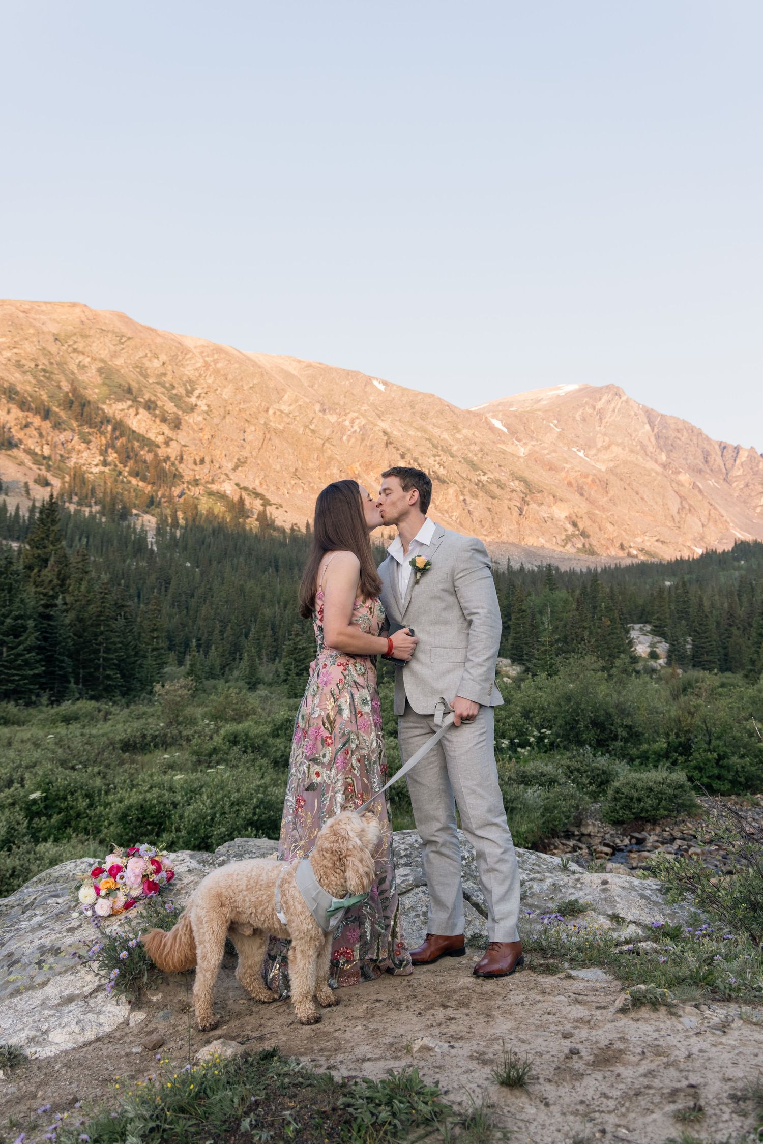 A couple shares a romantic moment with their goldendoodle dog in front of majestic mountain peaks at sunset.