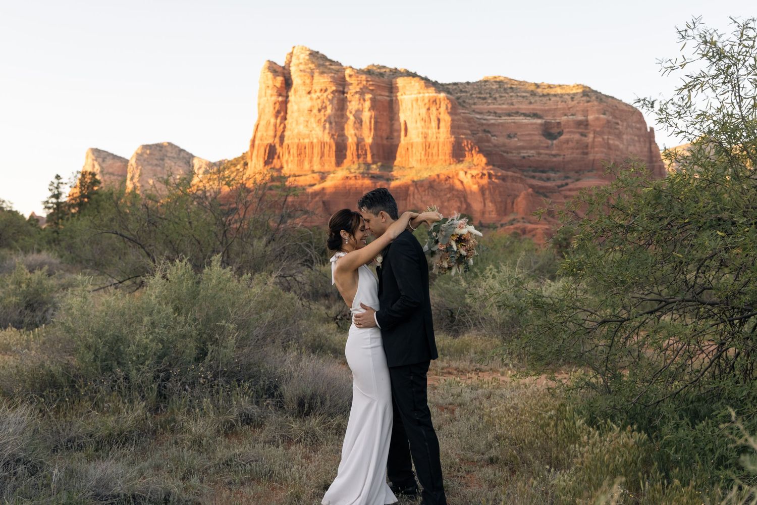 Couple in formal attire embraces while dancing against stunning red rock formations in Sedona at sunset.