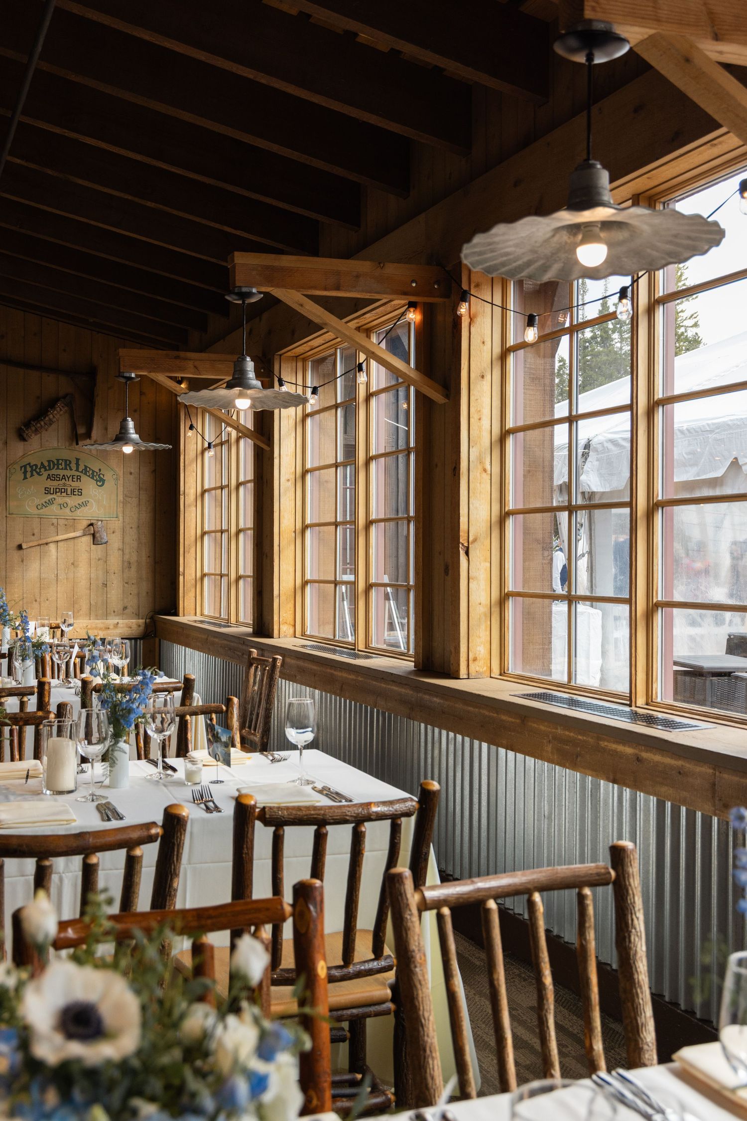 Rustic wedding reception venue with wooden chairs, set tables, large windows, and vintage pendant lights.