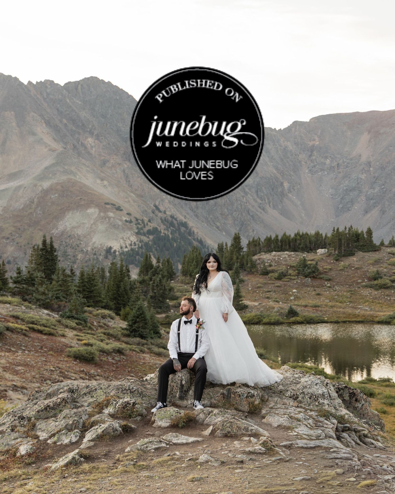Couple in wedding attire poses on mountain rocks with alpine lake and peaks, featured on Junebug Weddings.
