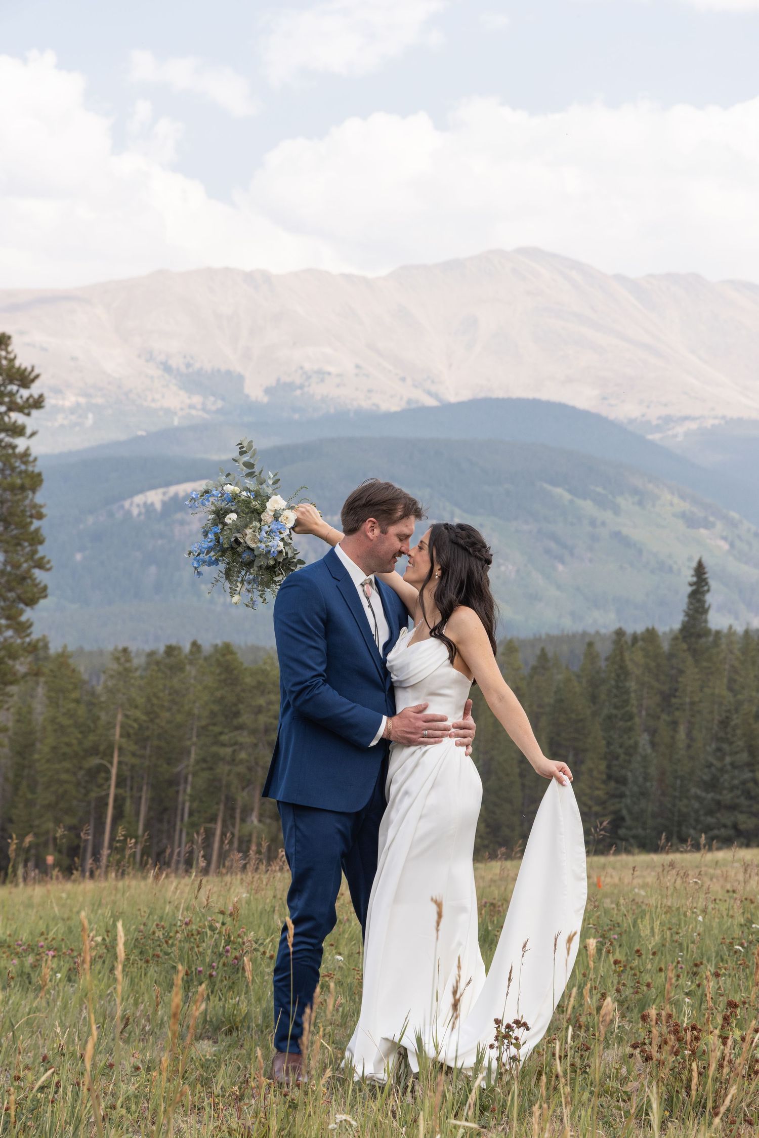 Bride and groom share a kiss in a mountain meadow, with evergreen trees and Rocky Mountain peaks in the background.