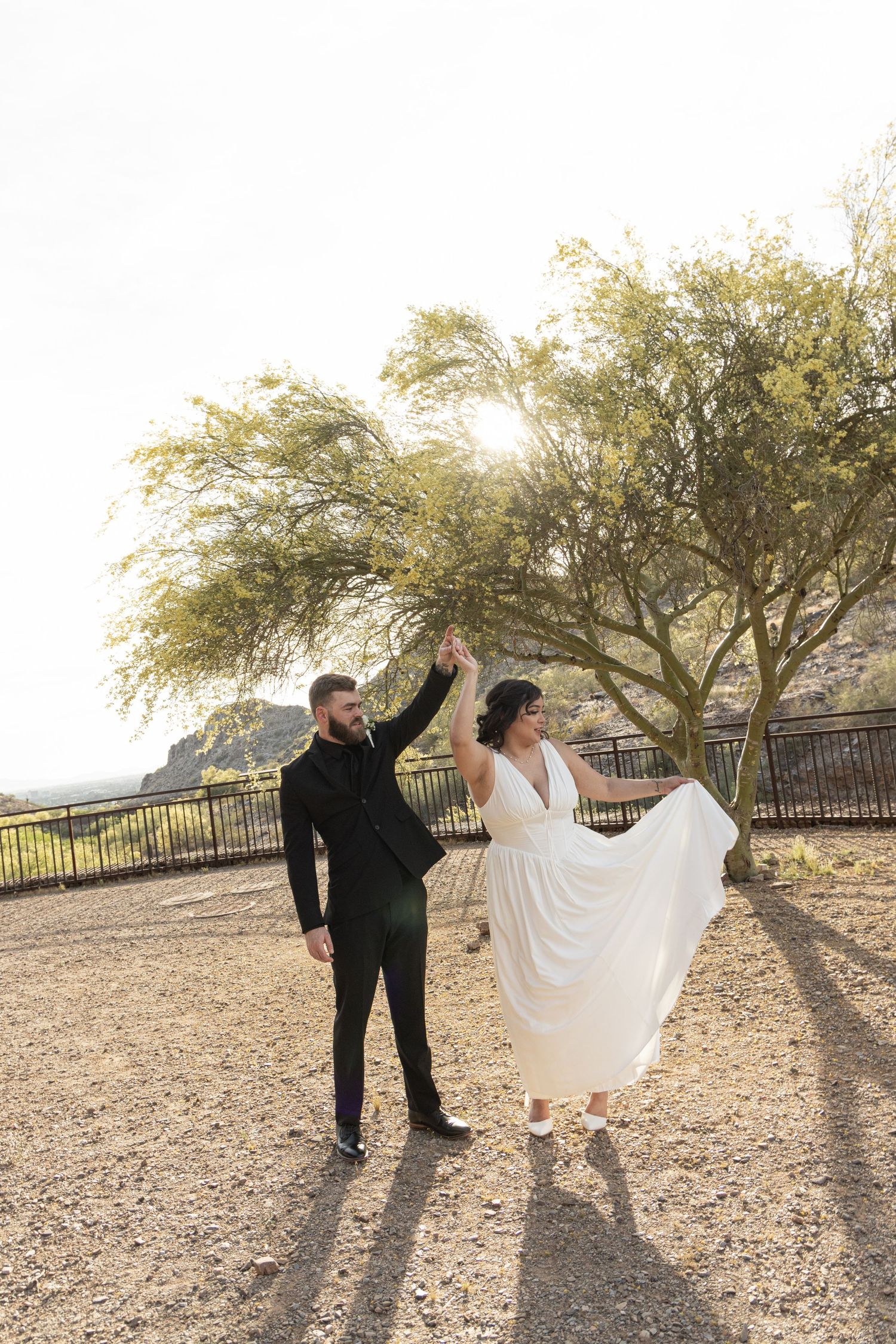 A couple in wedding attire dances and spins together on a desert path during golden hour.
