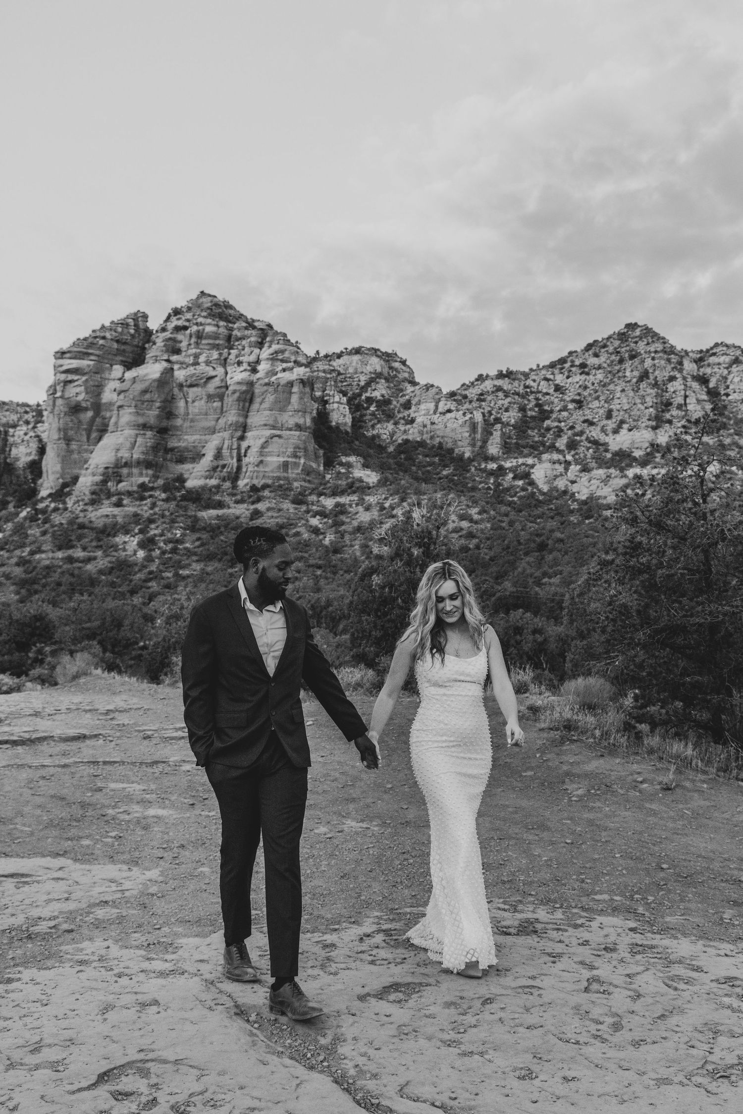 A couple in wedding attire walks together across red rock formations in Sedona during a scenic desert photo session.