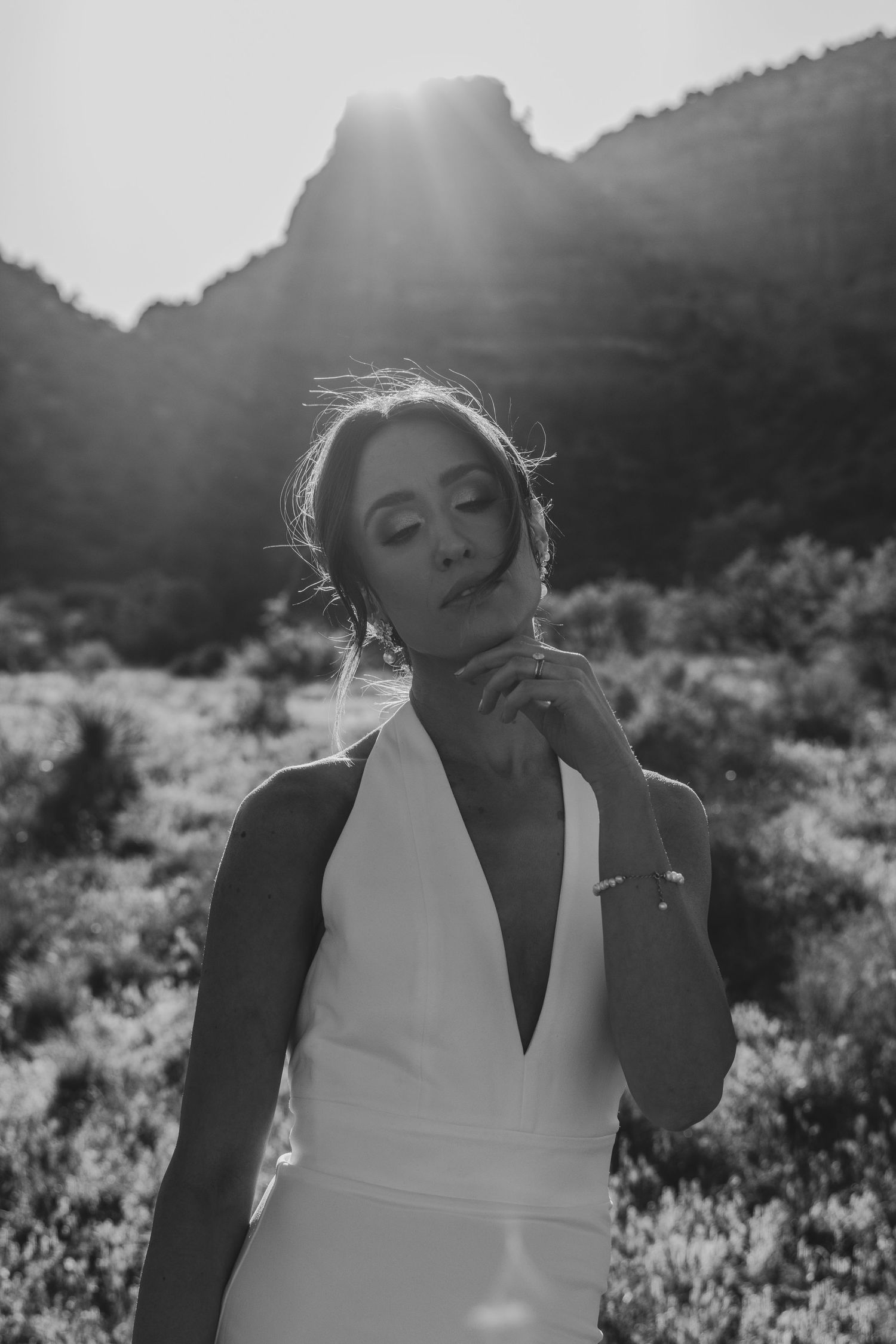 Black and white photo of someone in a white sleeveless dress posing outdoors with sunlight and mountains in the background.