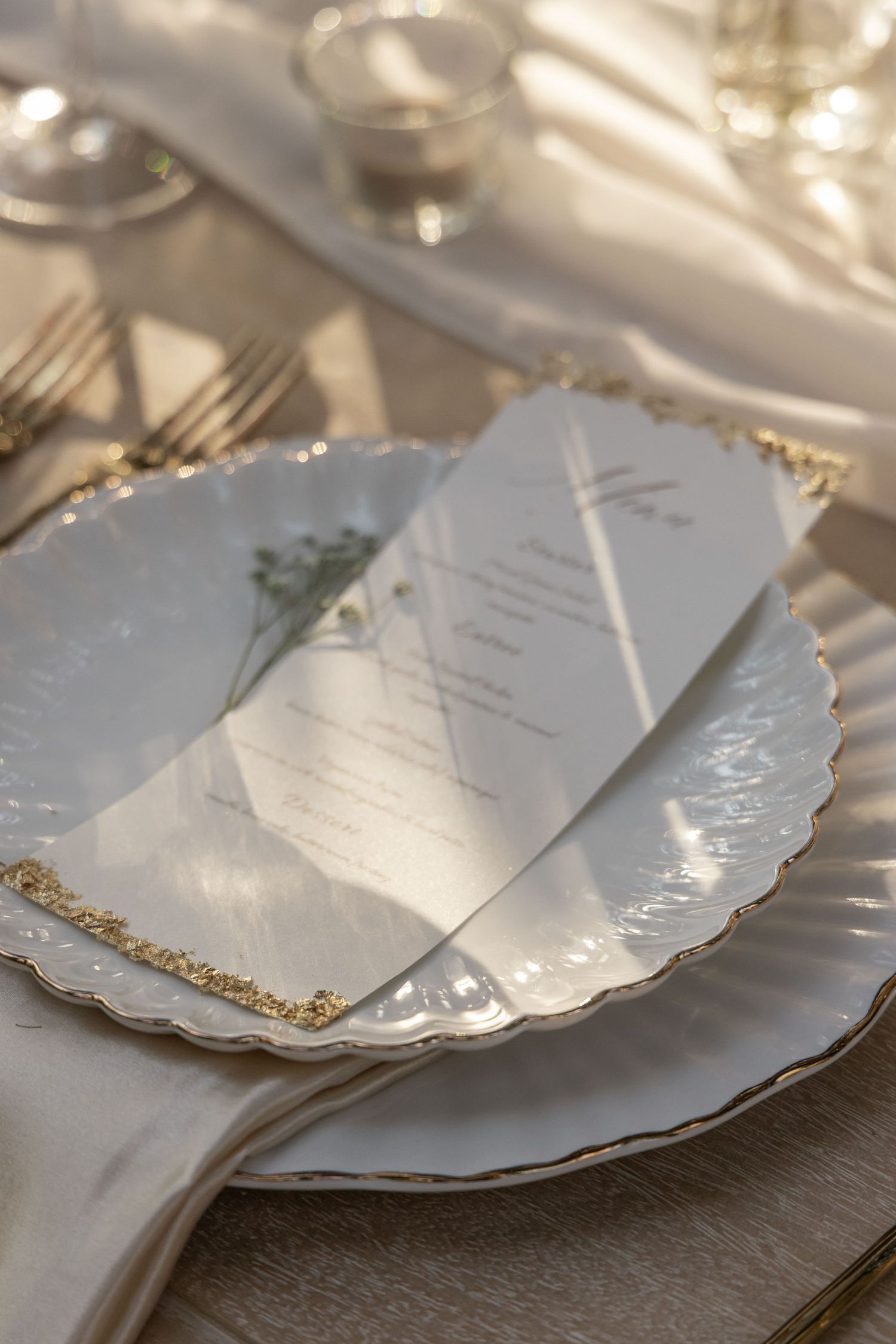 Elegant wedding place setting with gold-rimmed white charger plate, menu card, and crystal glassware in soft natural light.