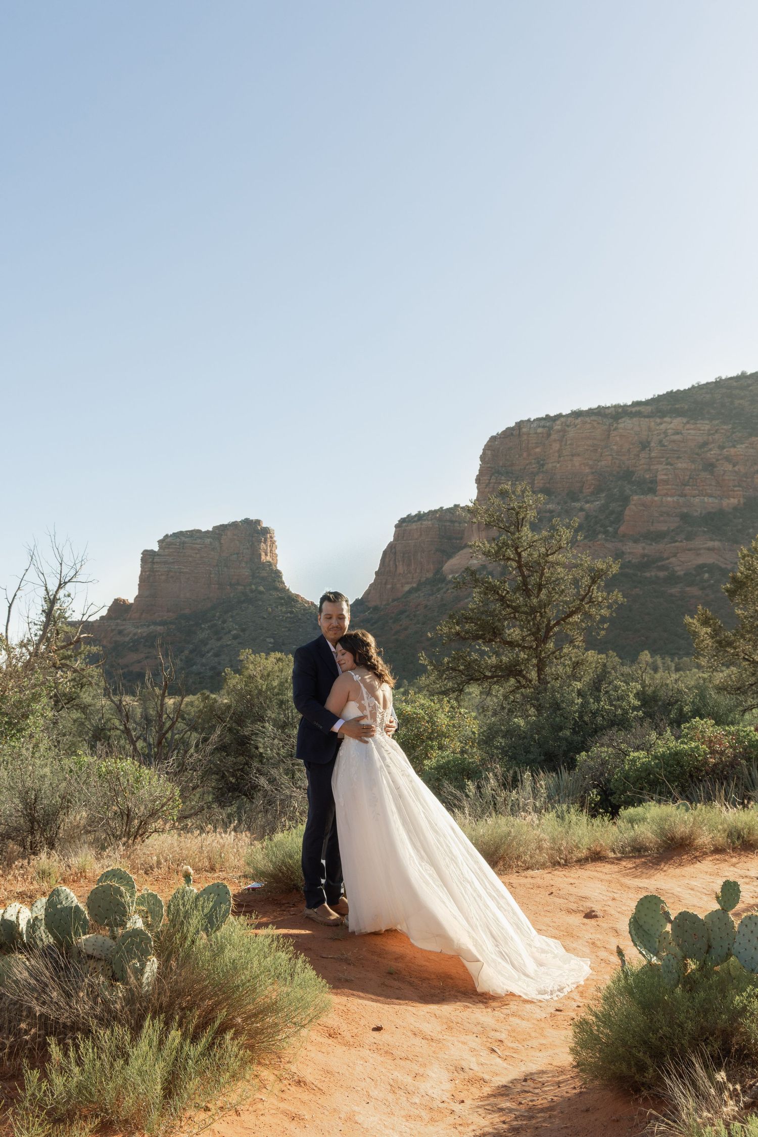 Bride and groom embrace on red dirt path in Sedona, Arizona, with iconic red rock buttes and desert landscape behind them.