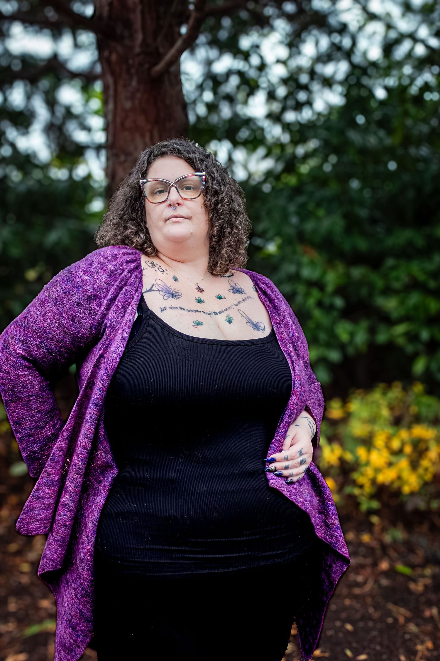 Person wearing a purple cardigan and black top poses outdoors with yellow flowers and greenery in background.