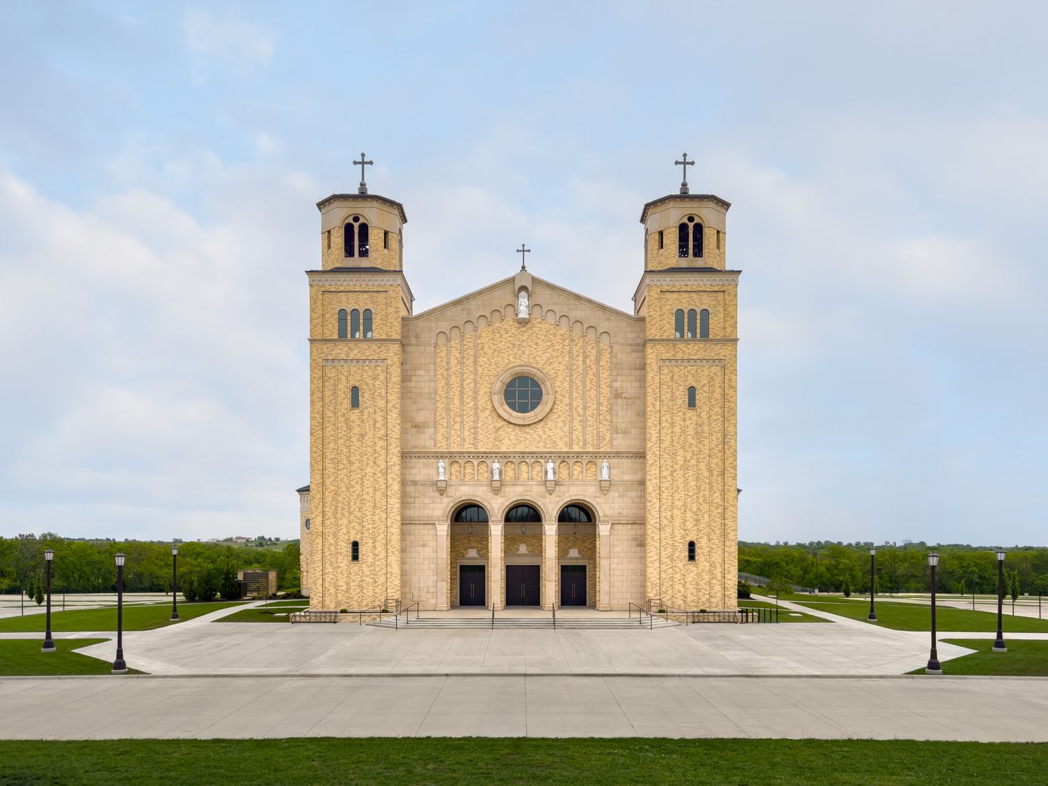 Symmetrical Romanesque Revival church with twin bell towers, arched entrance, rose window, and manicured grounds under a clear sky.