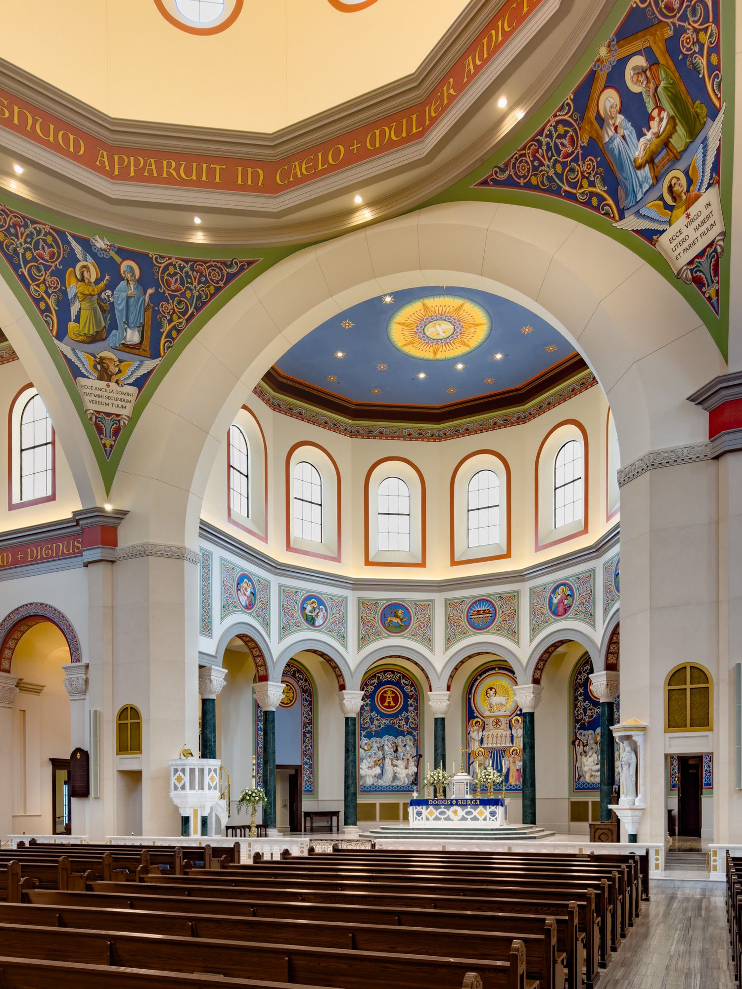 Ornate Catholic church interior with colorful frescoes, arched ceilings, wooden pews, and a decorated altar sanctuary.