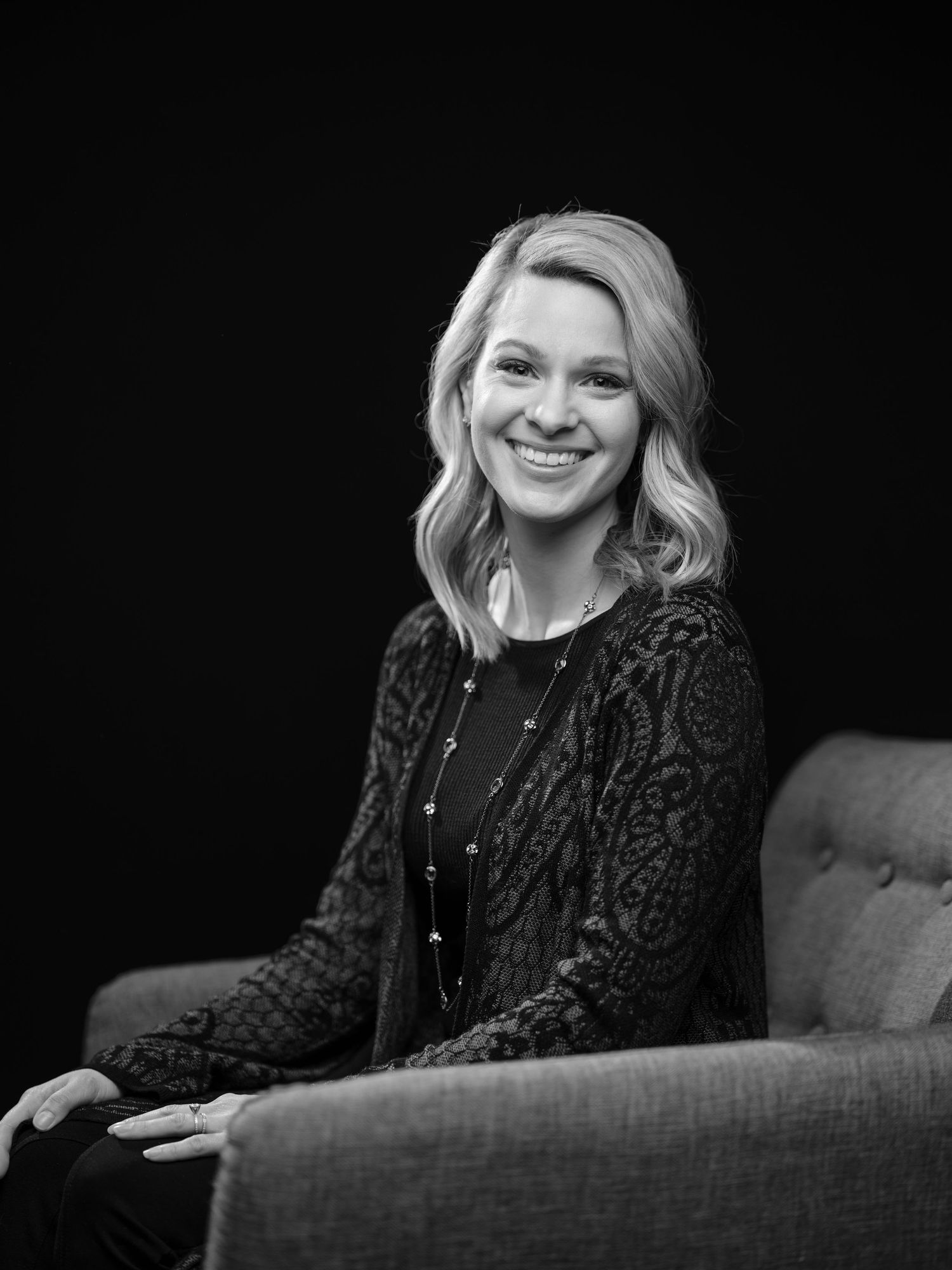 Black and white portrait of a person wearing a dark lace sweater sitting on a couch, smiling warmly at the camera.