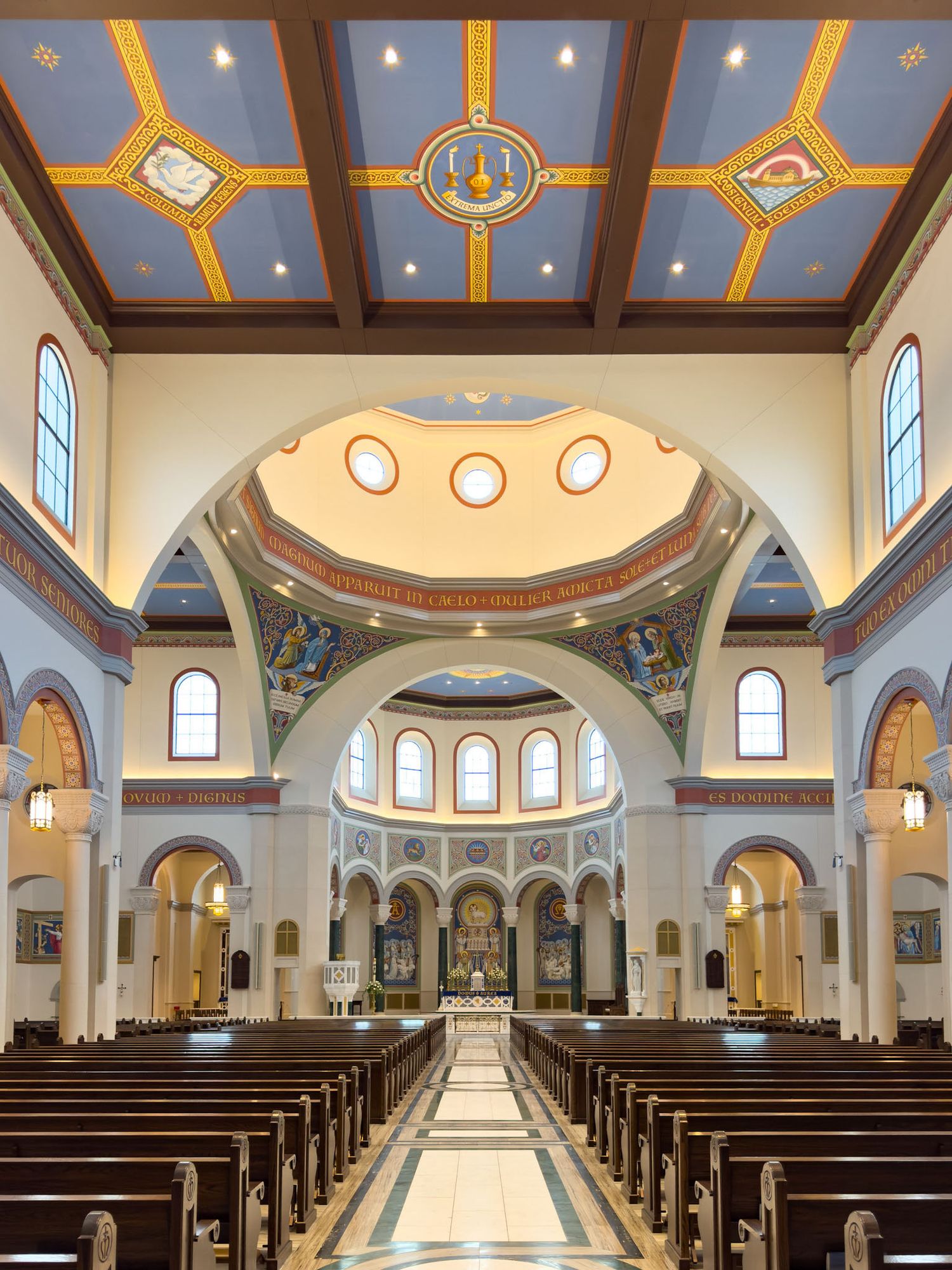 Grand Catholic church interior with ornate painted ceiling, arched nave, marble floors, and wooden pews leading to decorated altar.