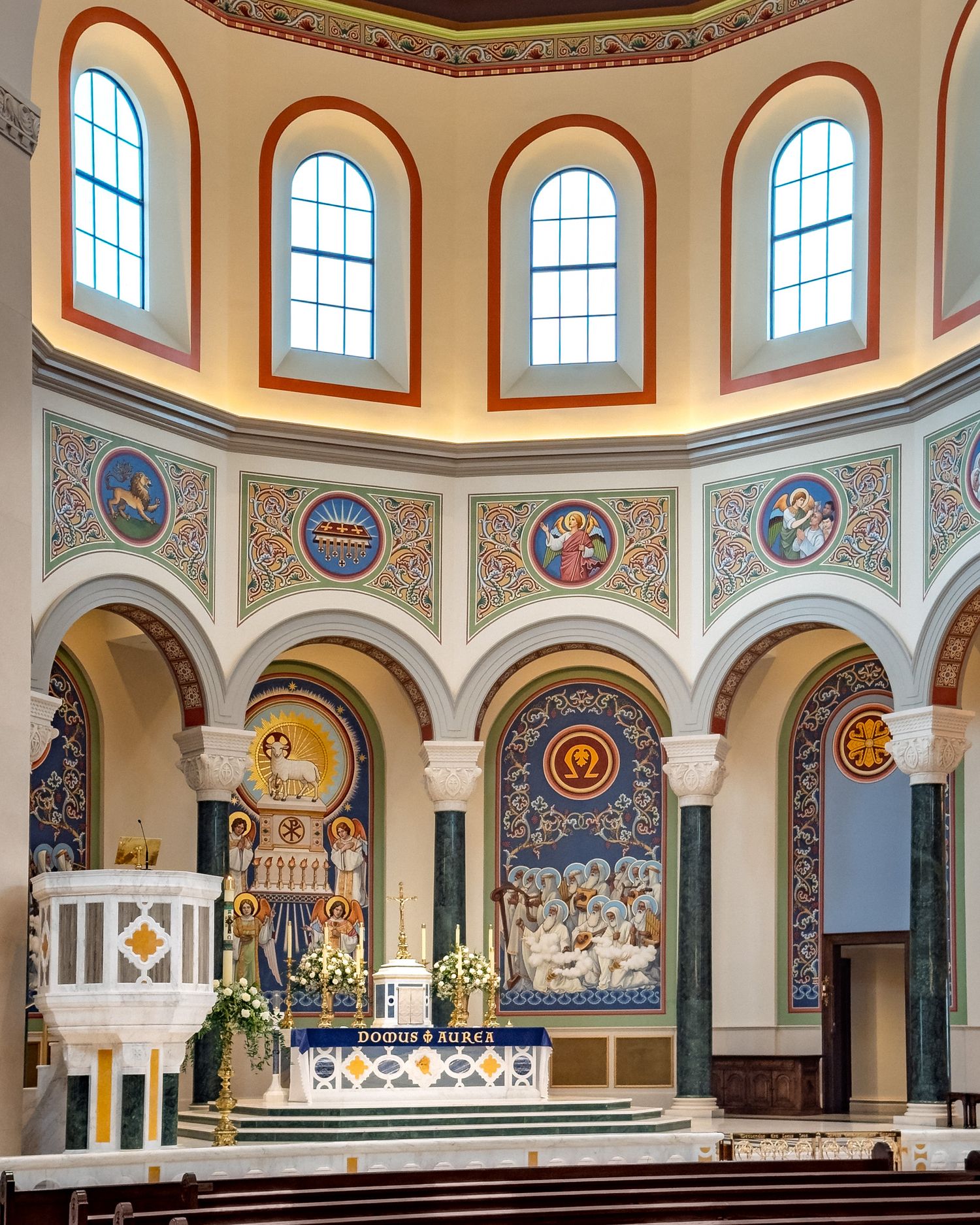 Ornate Catholic church interior with colorful mosaic murals, arched windows, and decorated altar under a domed ceiling.