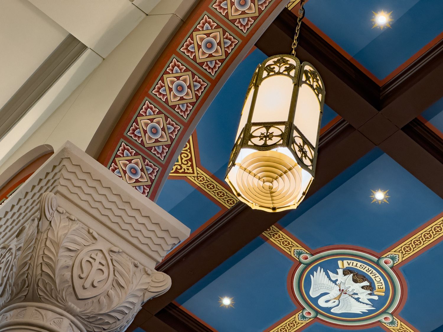 Ornate ceiling with colorful medallions, decorative moldings, a hanging lantern, and an intricately carved stone column capital.