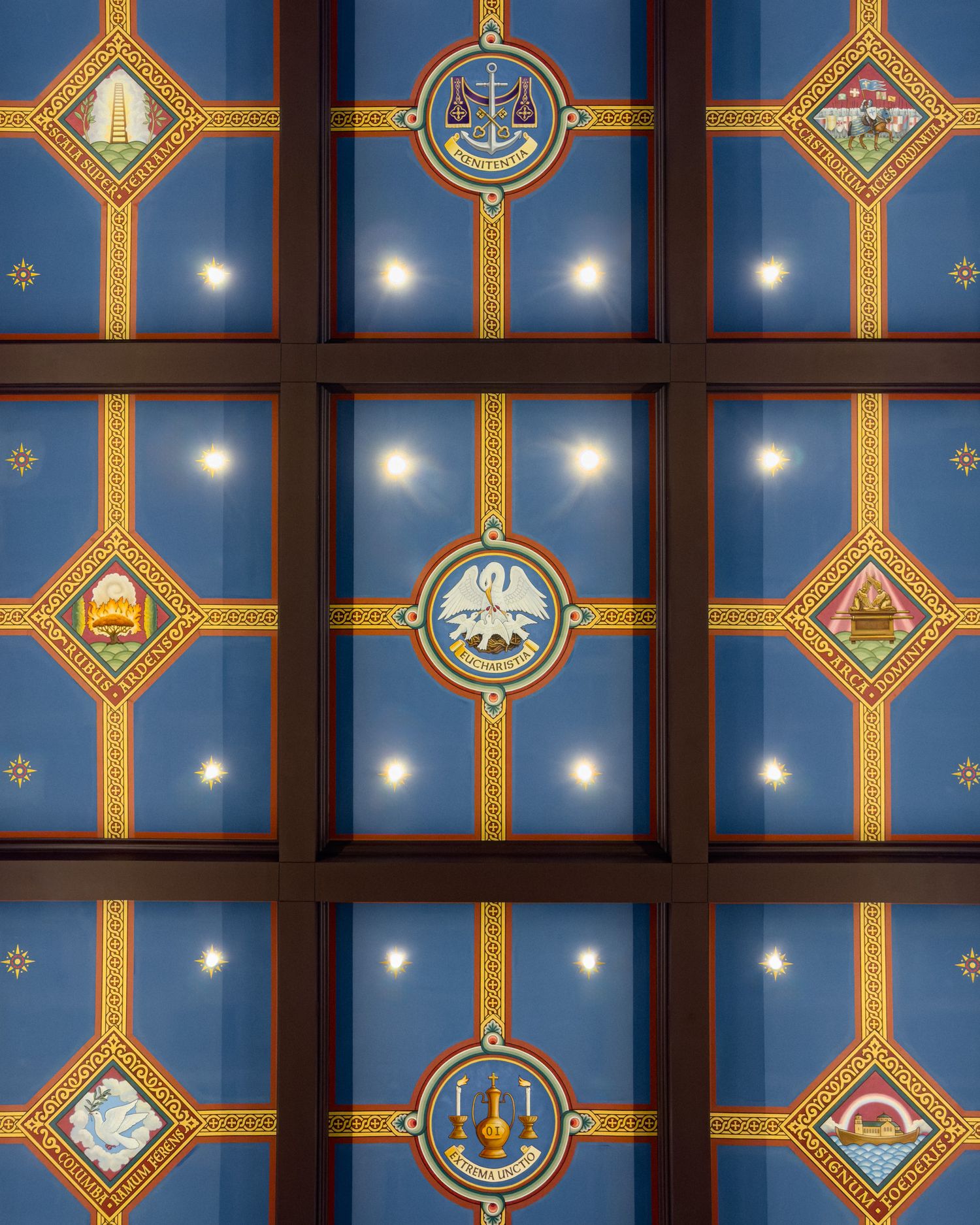 Ornate blue ceiling with gold-framed panels featuring colorful medallions and emblems, illuminated by recessed lights.