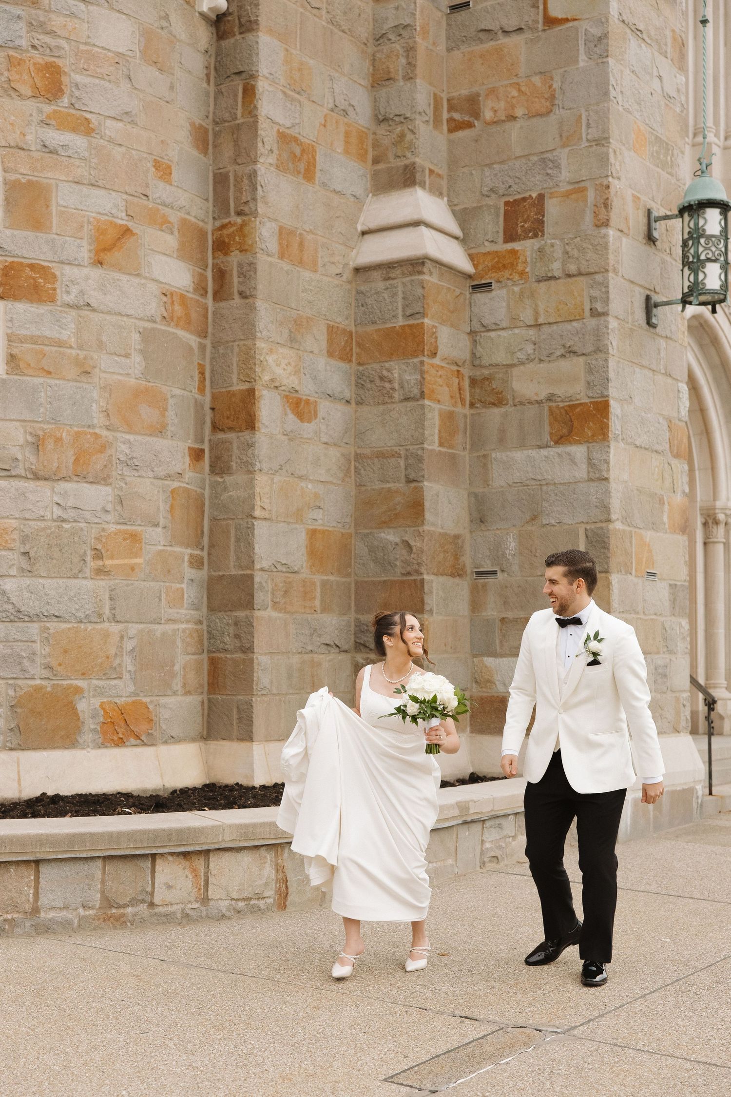 A bride and groom walk together outside a stone church building with elegant architecture.