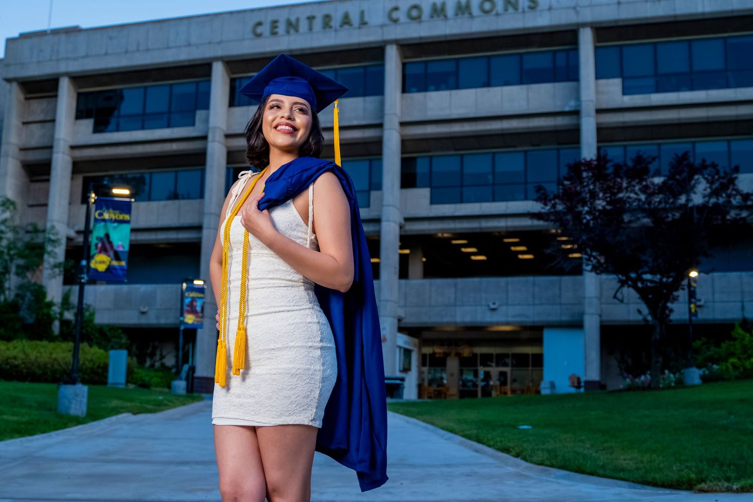 Graduate in white dress and blue cap and gown poses in front of Central Commons building. santa clarita.