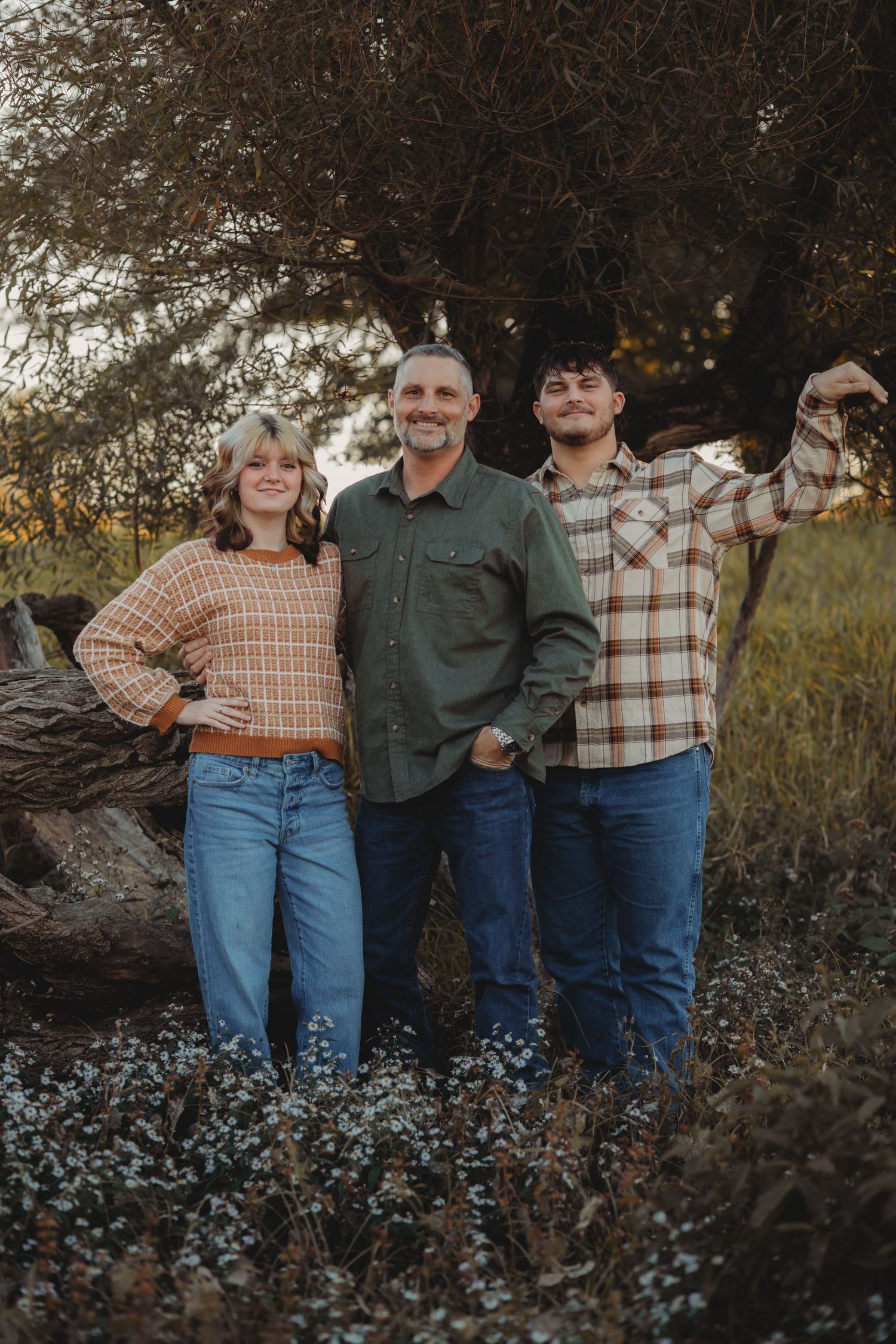 Family of three smiling outdoors near a large tree in a field, wearing casual fall outfits at golden hour.