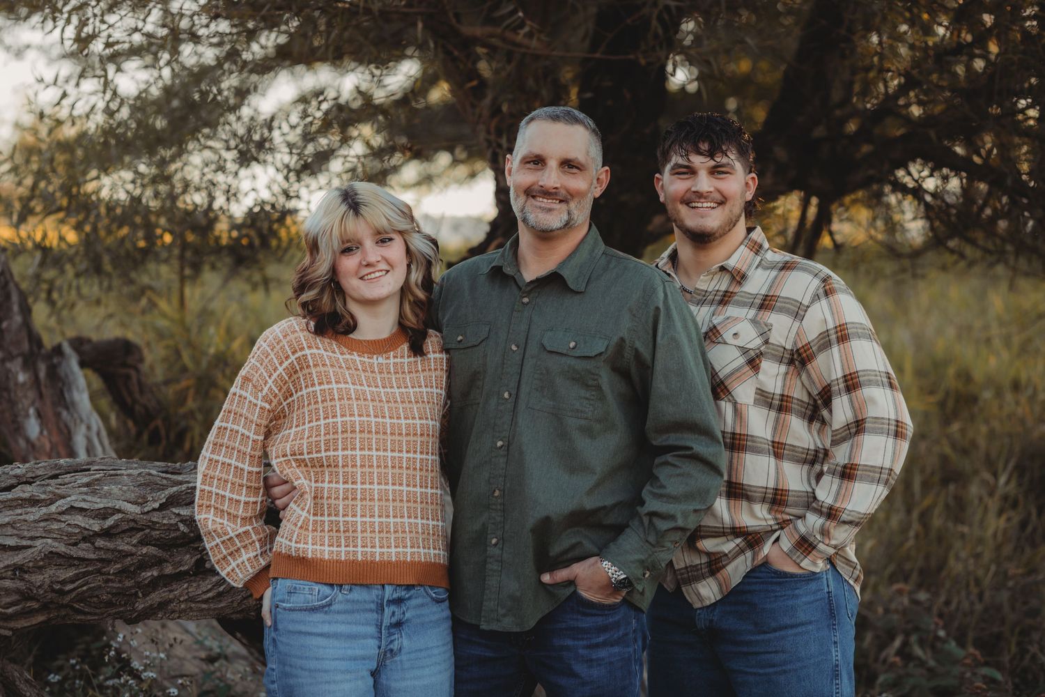 Family of three smiling outdoors in plaid and green shirts, posing in a natural setting with trees and golden light.
