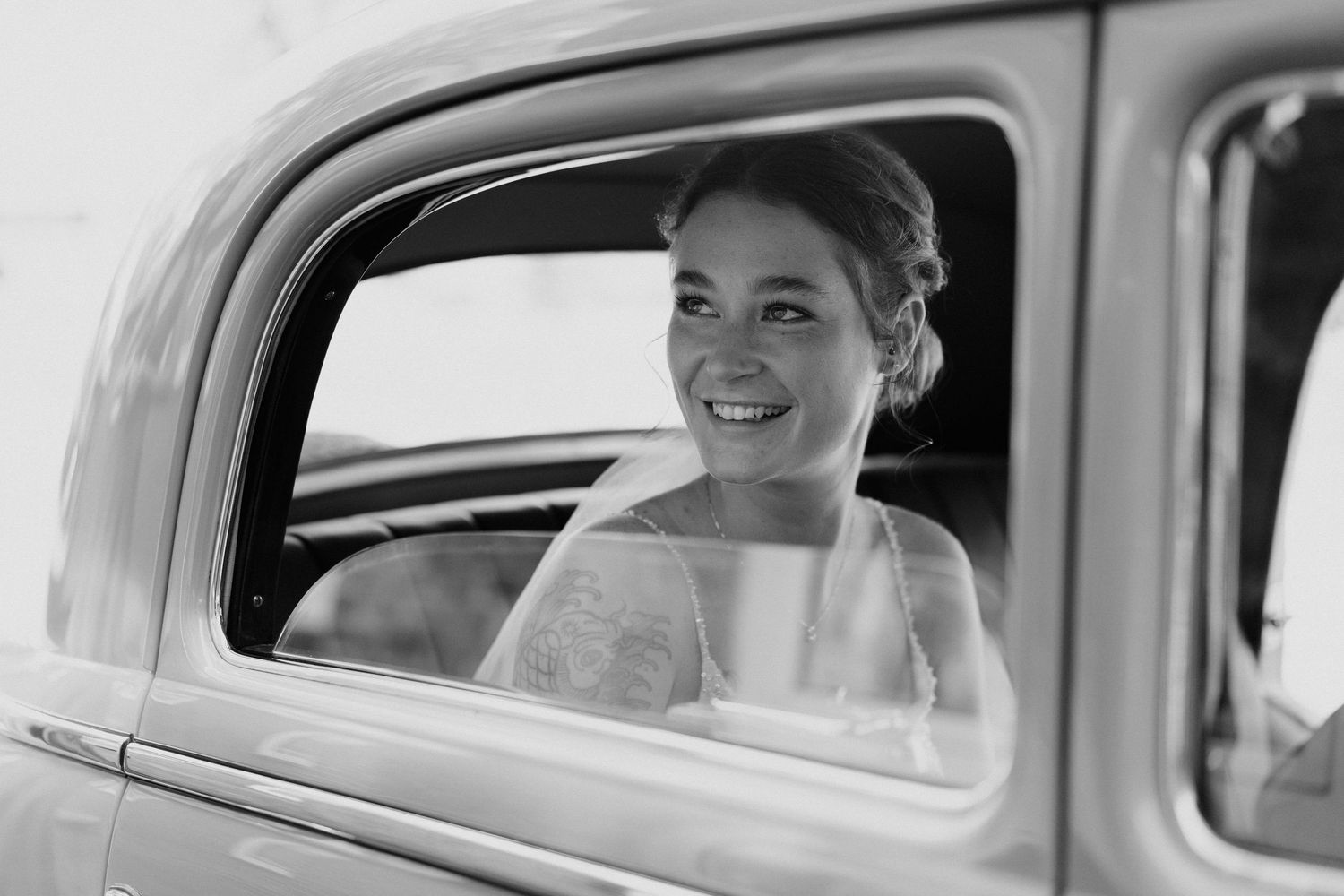 Radiant bride smiling through vintage car window in classic black and white wedding portrait.